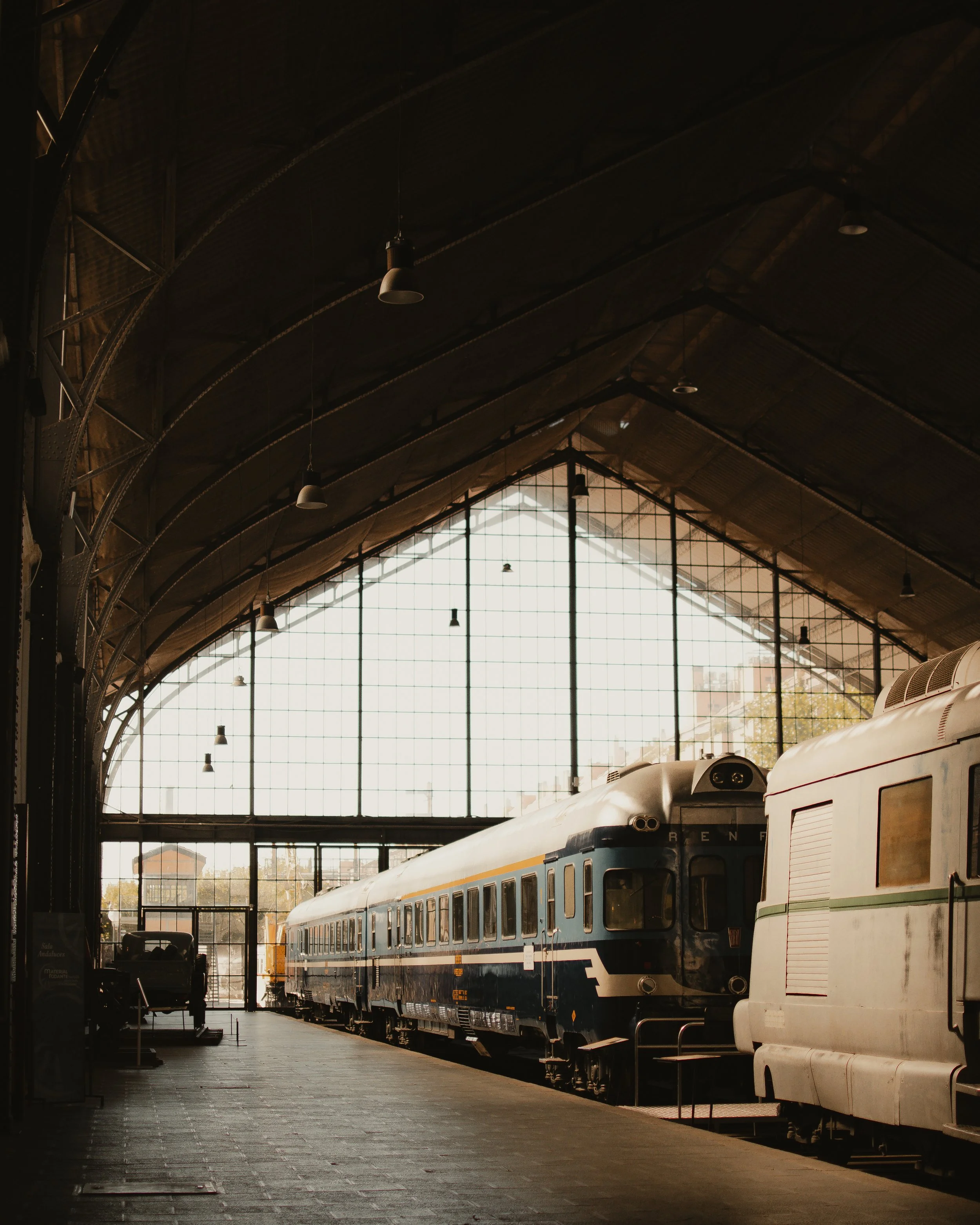 Interior de una estación de tren histórica en Madrid con trenes antiguos y un techo de metal. La luz entra a través de grandes ventanas de vidrio.