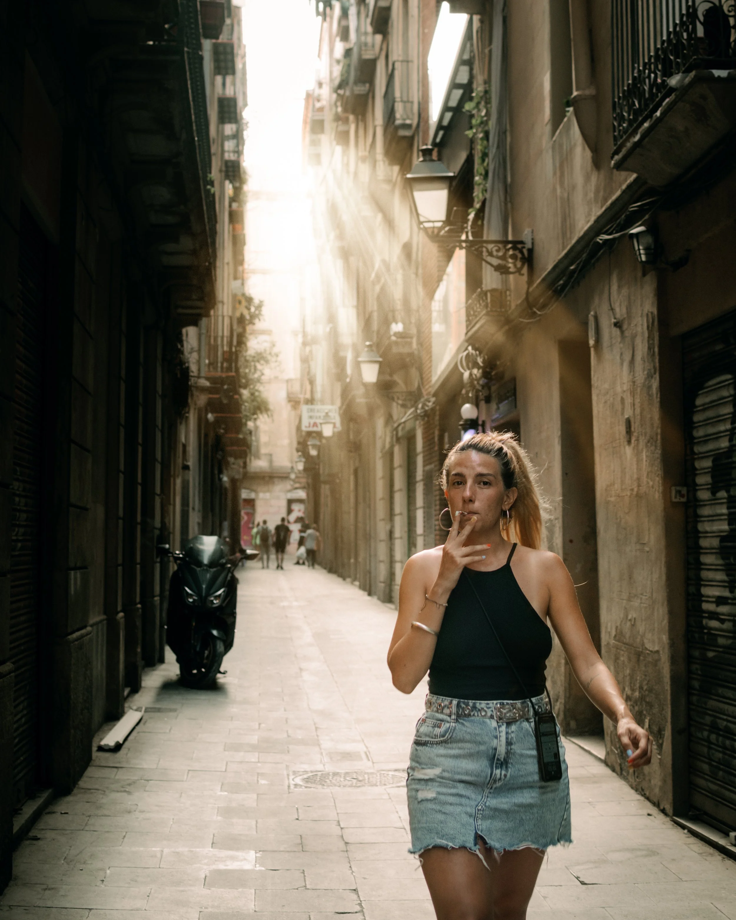 Una mujer caminando por una calle estrecha en Barcelona con edificios altos a los lados, luz del sol filtrándose entre los edificios. La mujer lleva una falda de mezclilla y una blusa sin mangas.