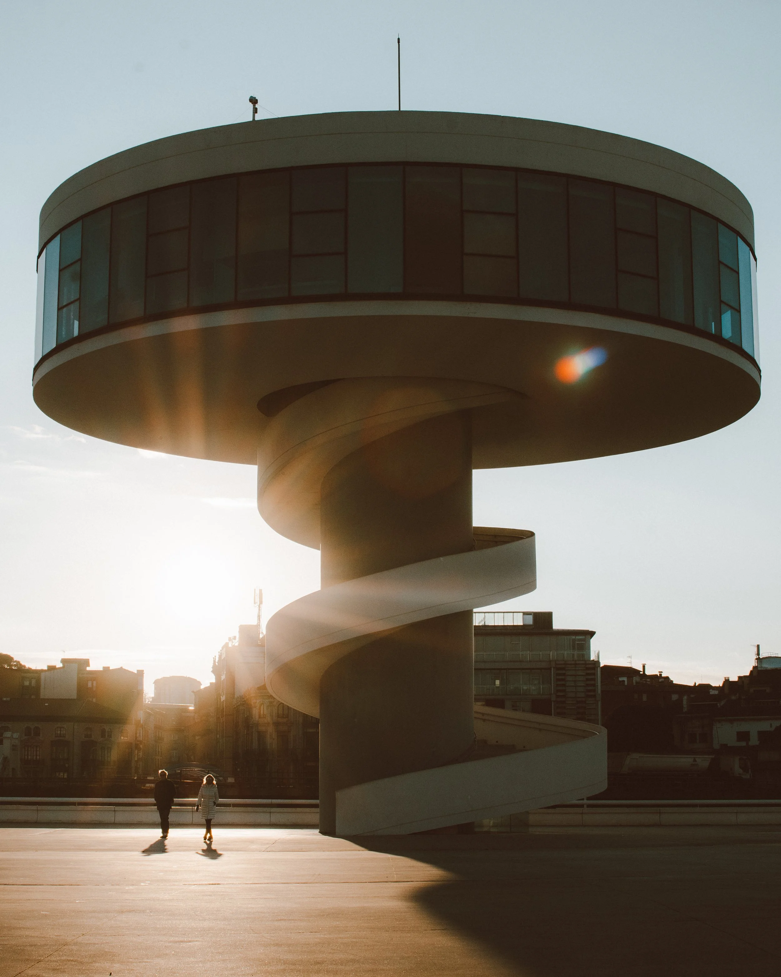 Vista de un edificio moderno con una estructura circular y una rampa espiral. Dos personas caminan en el área de una plaza, bajo la luz del atardecer. Centro Internacional Oscar Niemeyer, Avilés, Asturias.