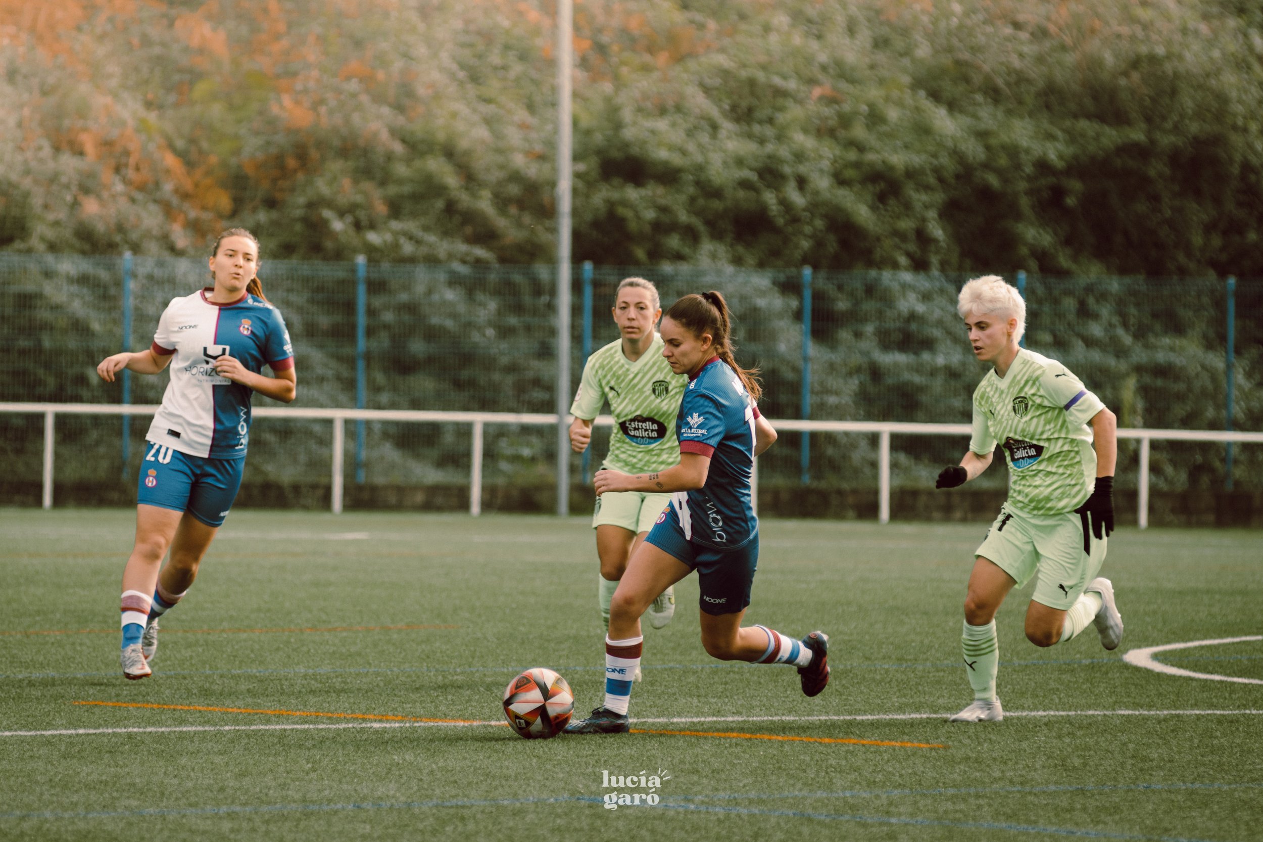 Jugadoras de fútbol femenil durante un partido, dos de ellas con uniforme azul y blanco y dos con uniforme verde claro, corriendo tras el balón en un campo de césped. Al fondo, una cerca y vegetación.