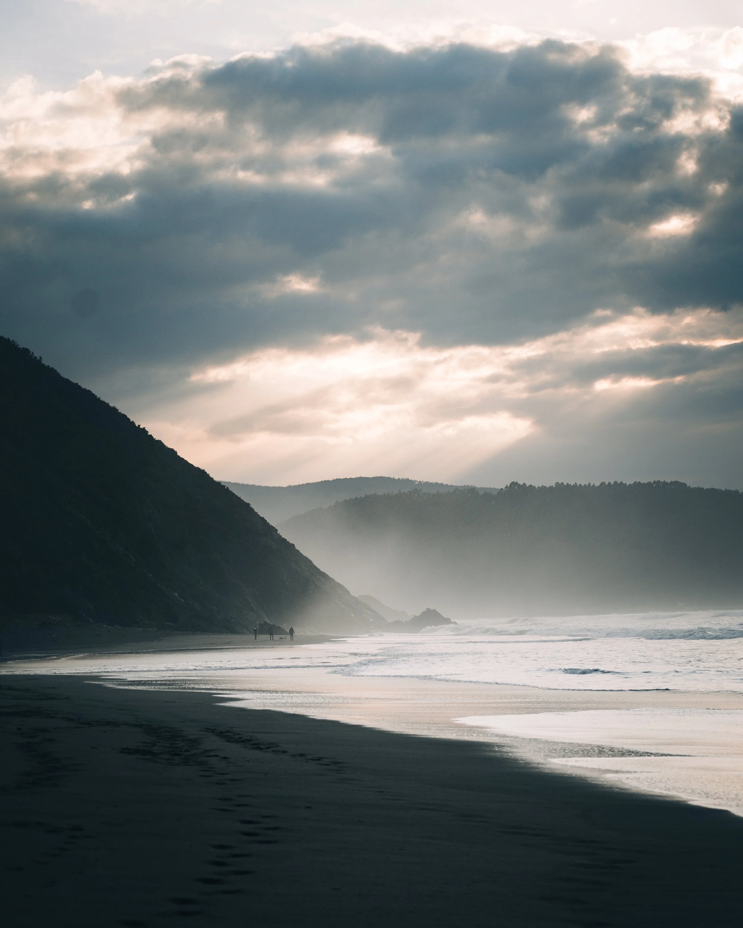 Playa al atardecer con montañas al fondo, nubes oscuras en el cielo y huellas en la arena.