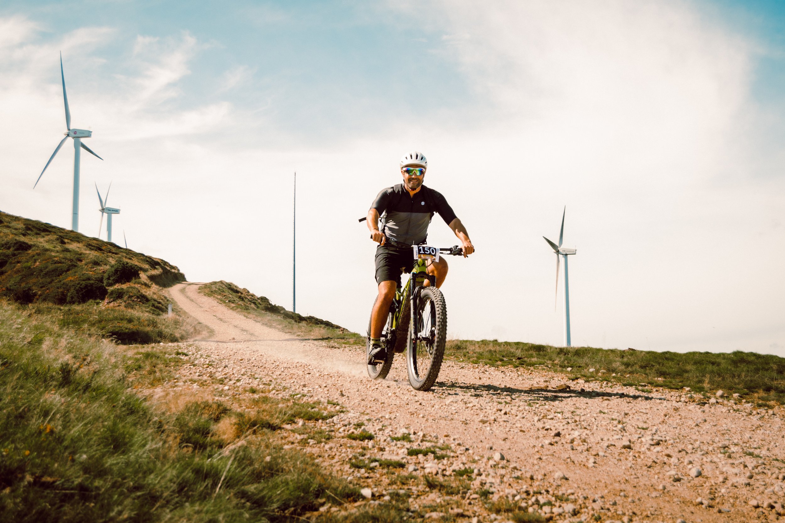 Ciclista en bicicleta de montaña en un camino de tierra cerca de turbinas eólicas en un paisaje rural.