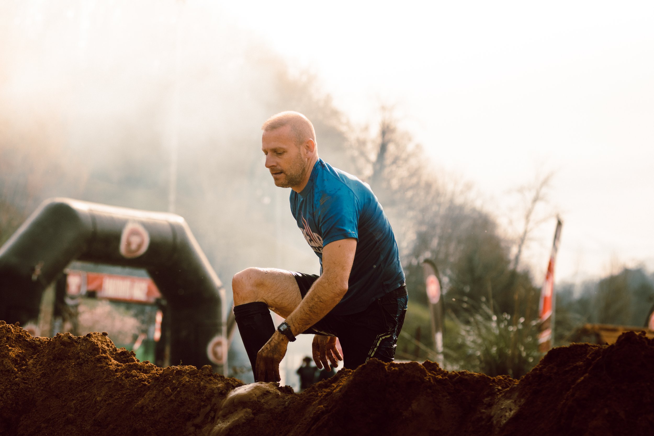 Un hombre participando en una carrera de obstáculos, subiendo un montículo de tierra, vestido con camiseta azul y pantalones cortos negros, en un entorno al aire libre.
