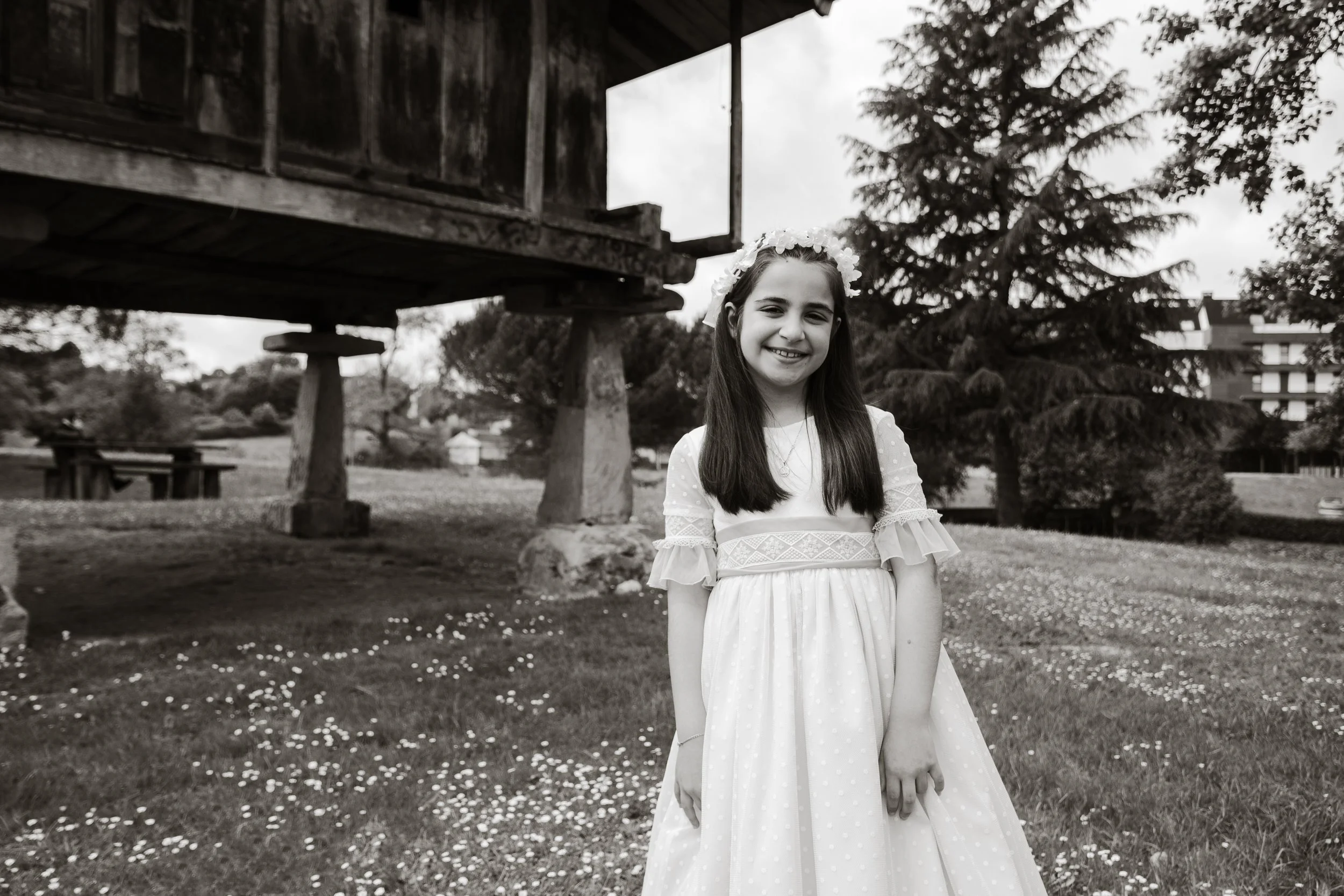 Niña con vestido de comunión blanco en un jardín, con un hórreo y árboles en el fondo. Imagen en blanco y negro. En Asturias