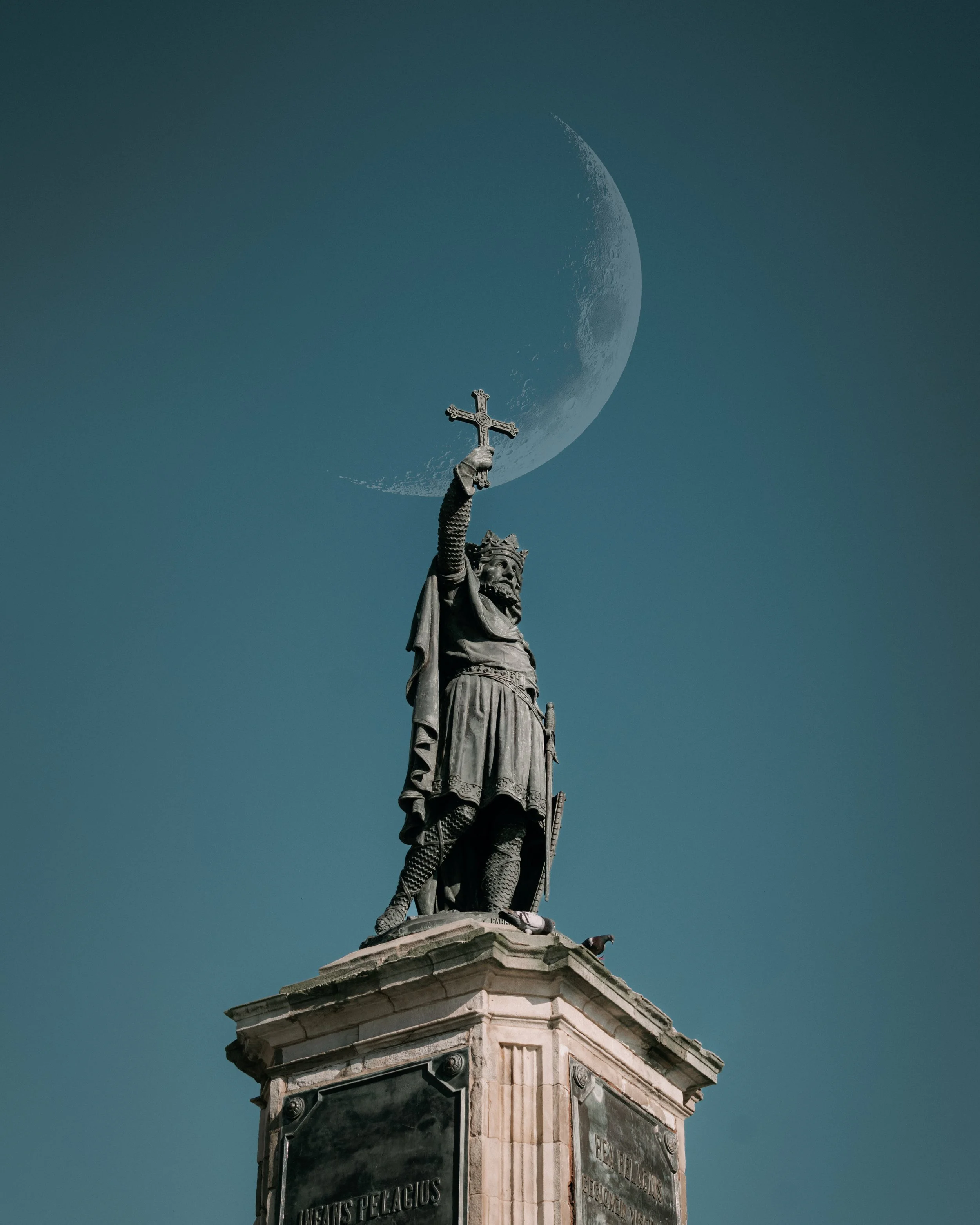 Estatua de un rey medieval en una base de piedra, sosteniendo una cruz con la luna creciente en el fondo, cielo azul.