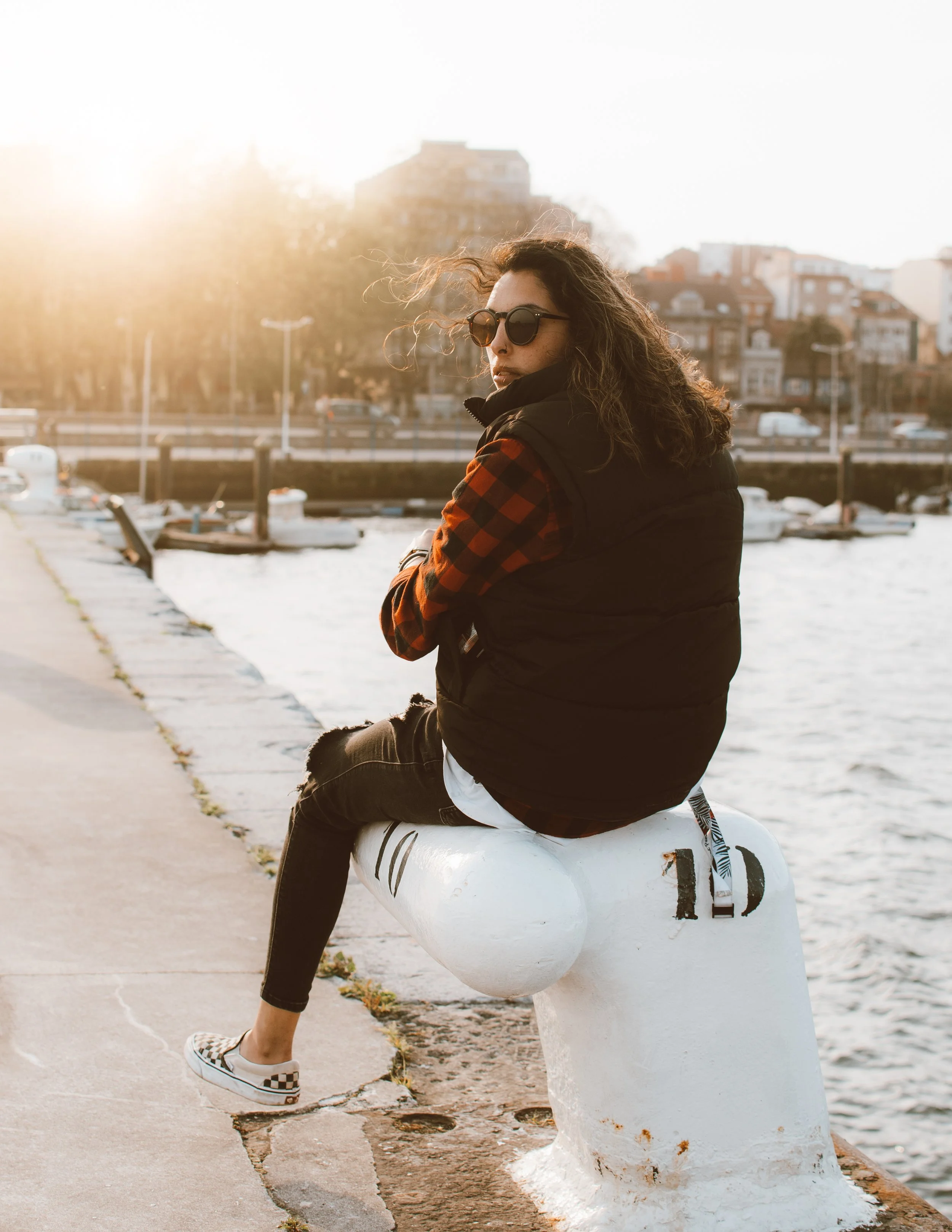 Persona sentada en un poste junto a un río o puerto al atardecer, usando gafas de sol y vestimenta casual.