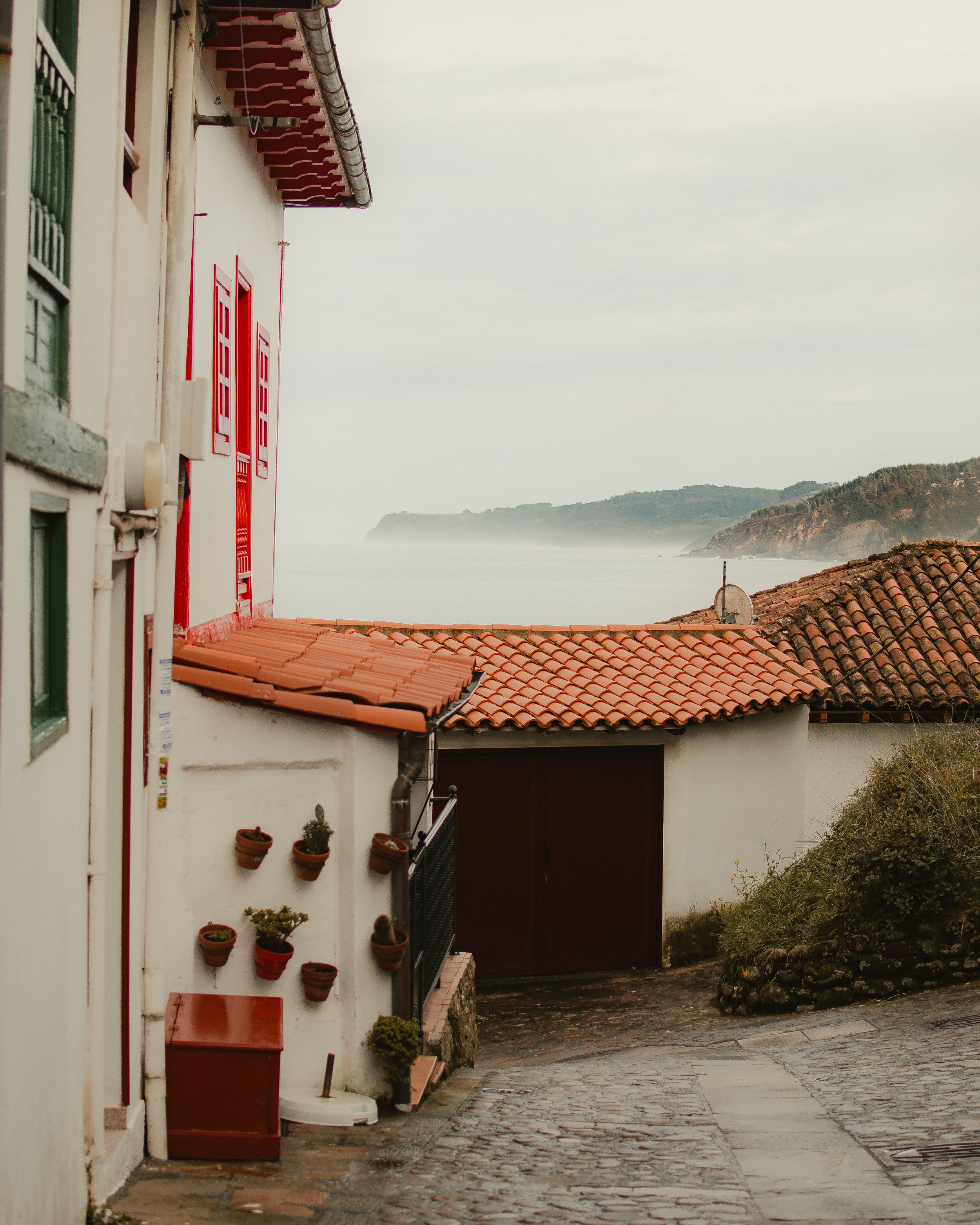 Calle estrecha en Tazones con casas de paredes blancas y techos de tejas rojas, vista al mar y colinas en el fondo, cielo nublado.