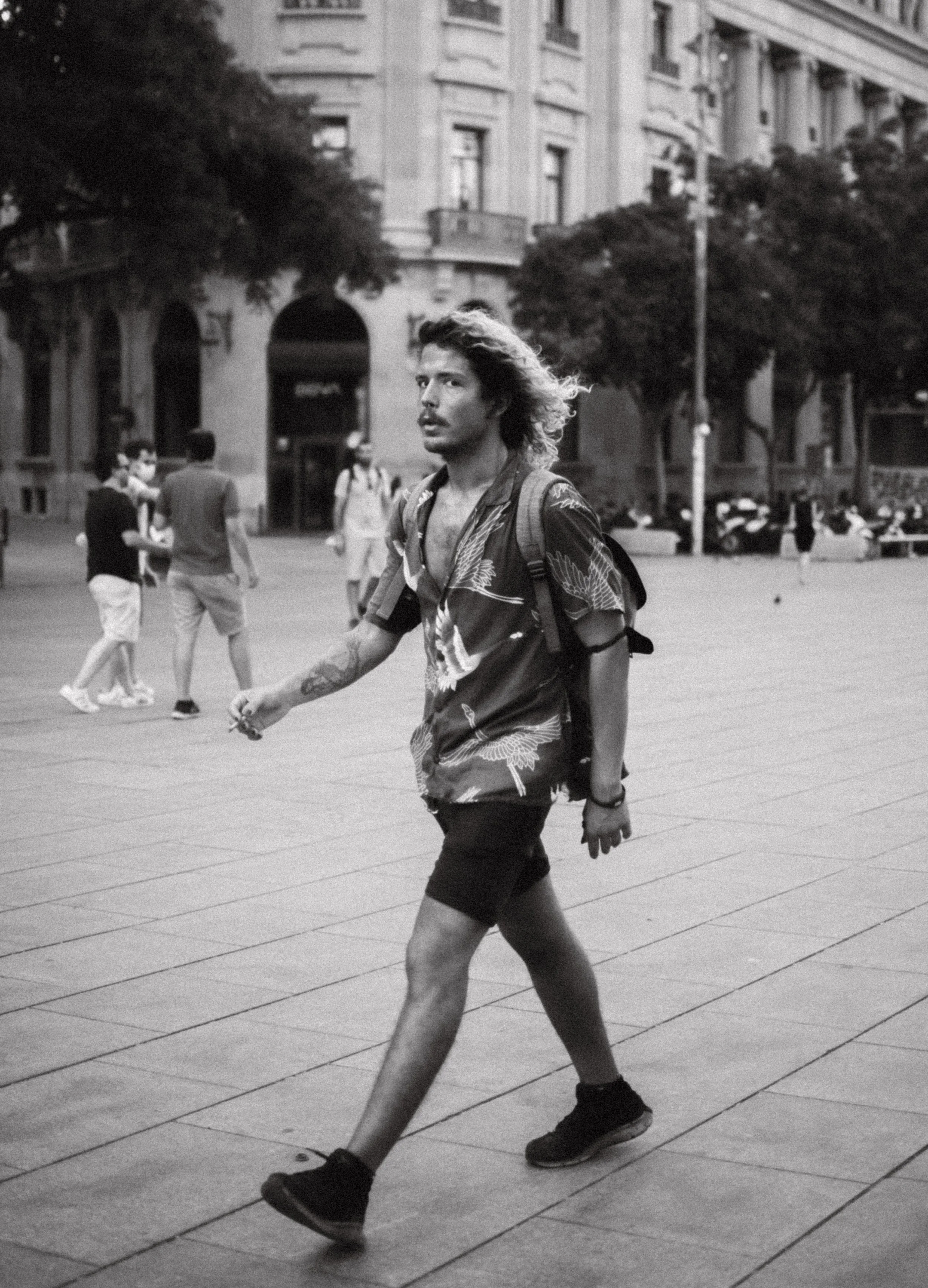 Hombre caminando en una plaza urbana, vistiendo camisa estampada y mochila, con edificios al fondo.
