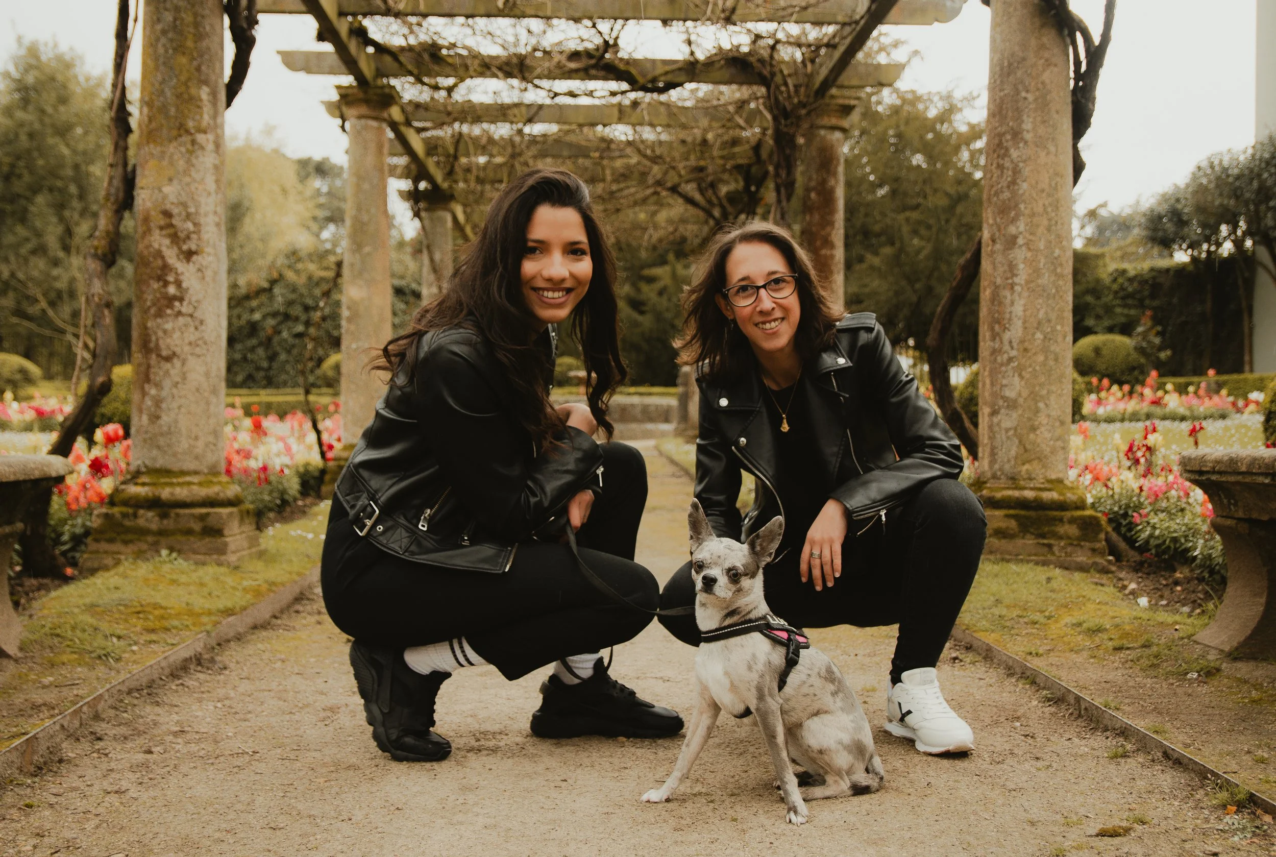 Dos personas sonriendo, agachadas junto a un perro, en un jardín con flores y columnas de piedra.