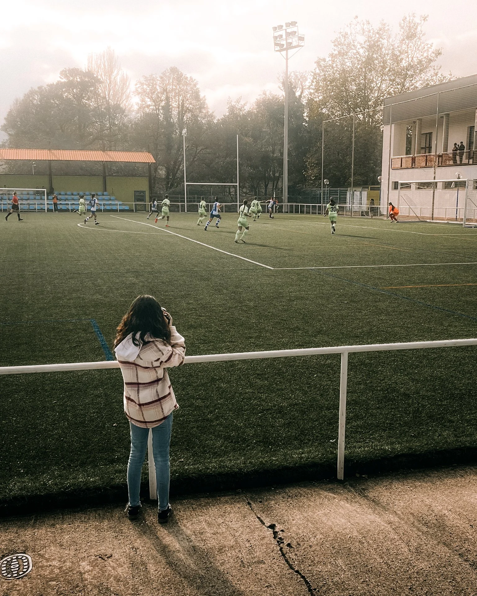 Lucía Garó, fotógrafa, observando un partido de fútbol en un campo al aire libre. Varios jugadores en acción, con camisetas verdes y blancas. Edificio y árboles al fondo. Clima nublado y luz suave.