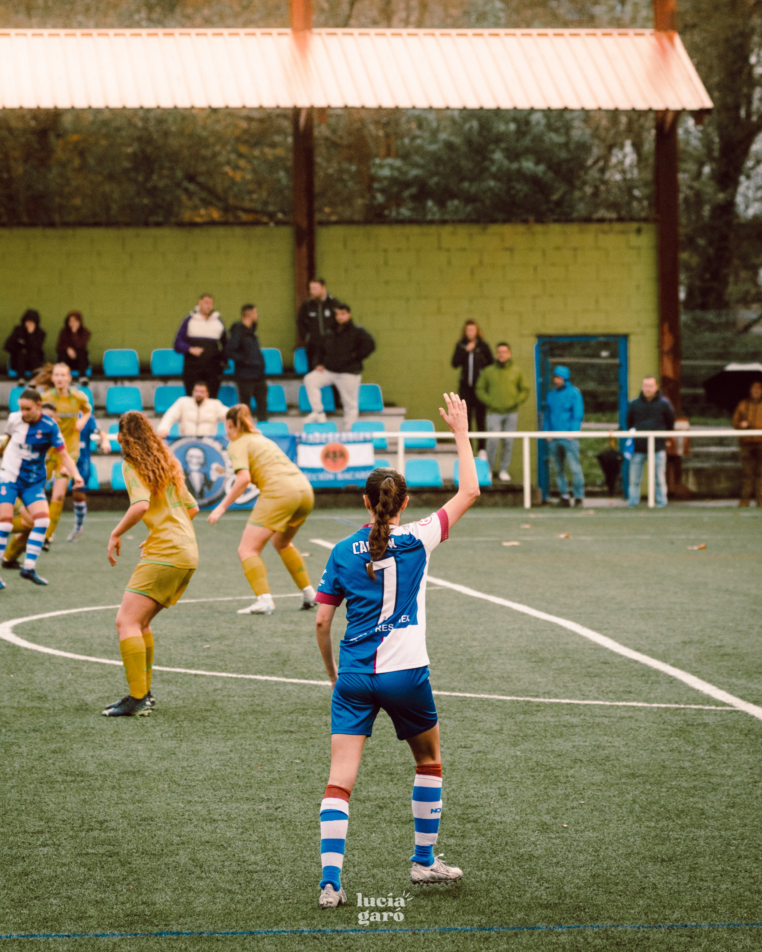 Jugadoras de fútbol en acción durante un partido en una cancha de pasto sintético, con espectadores al fondo.