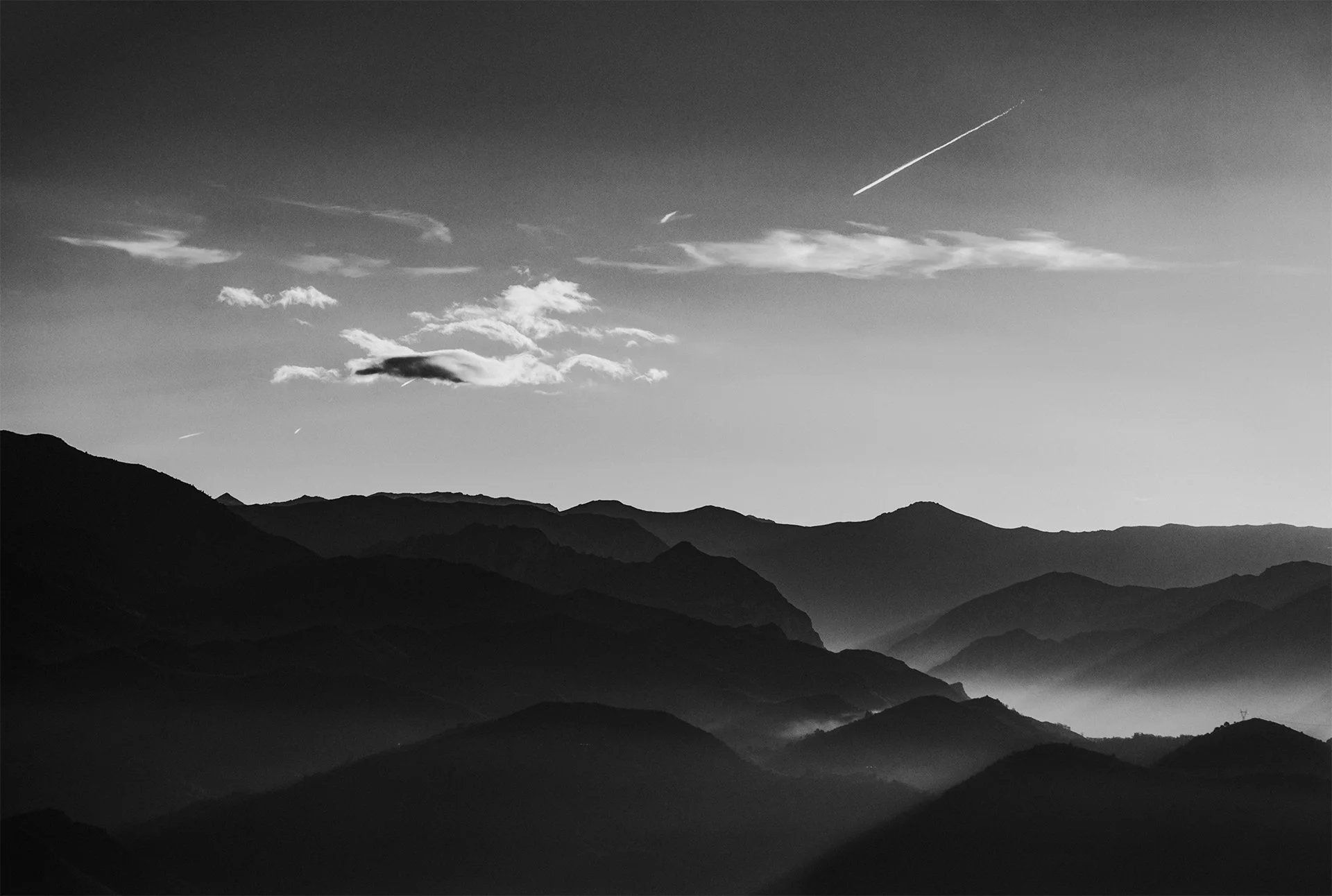 Paisaje de montañas en blanco y negro con nubes y estela de avión en el cielo.
