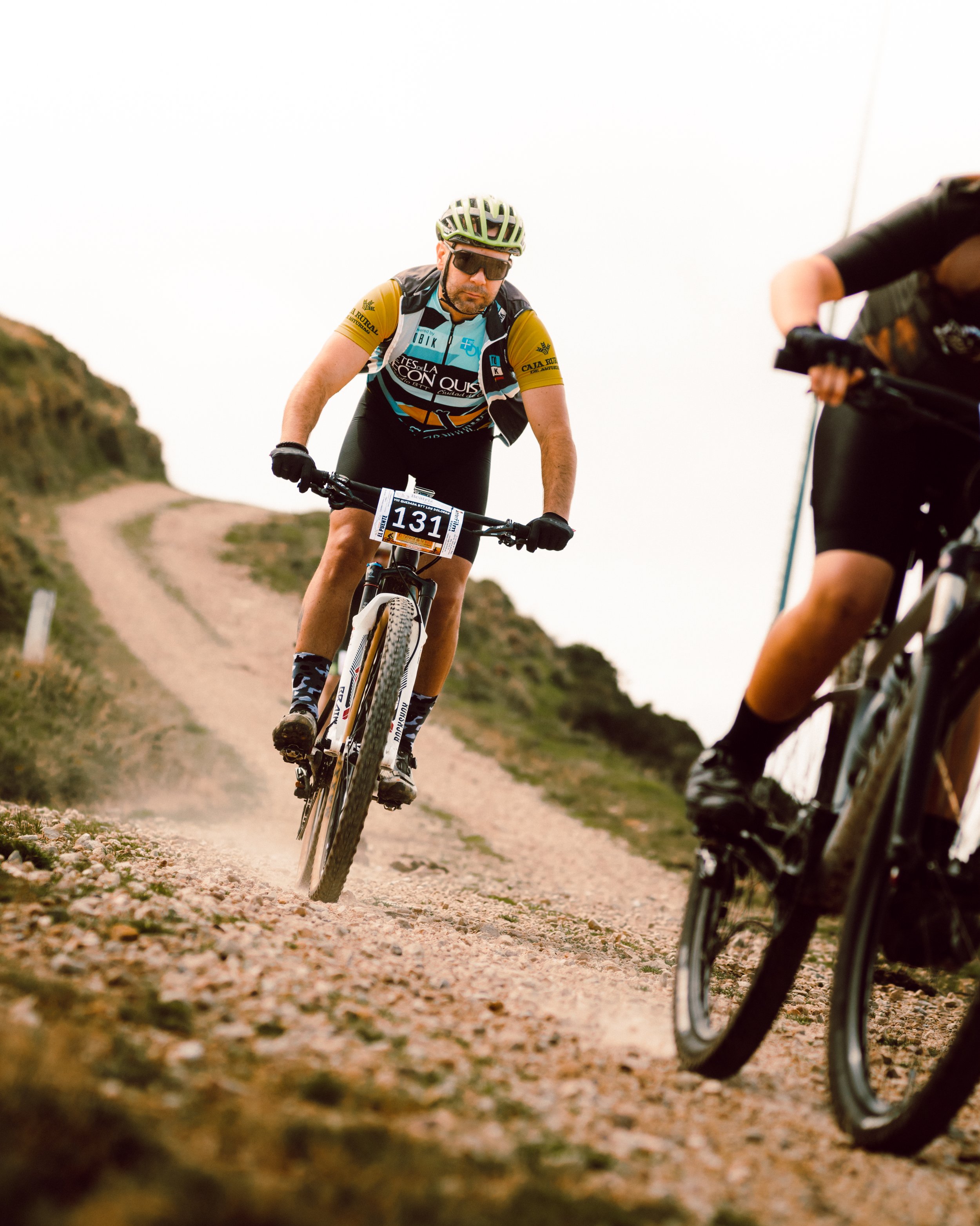 Dos ciclistas participando en una carrera de montaña en un camino de tierra; uno lleva un casco y gafas de sol.
