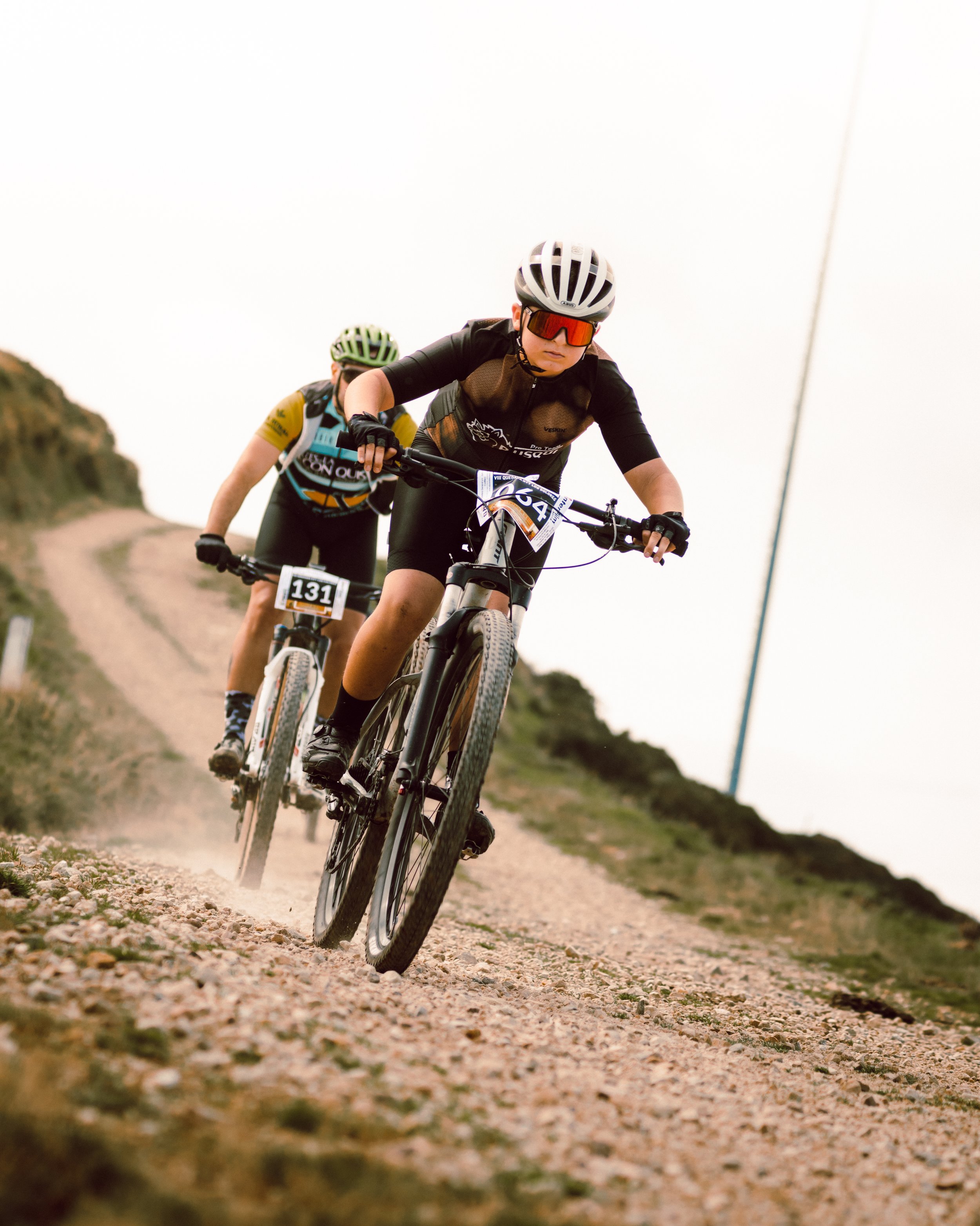Dos ciclistas en un camino de tierra compitiendo en una carrera de ciclismo de montaña, con cascos y gafas de protección.
