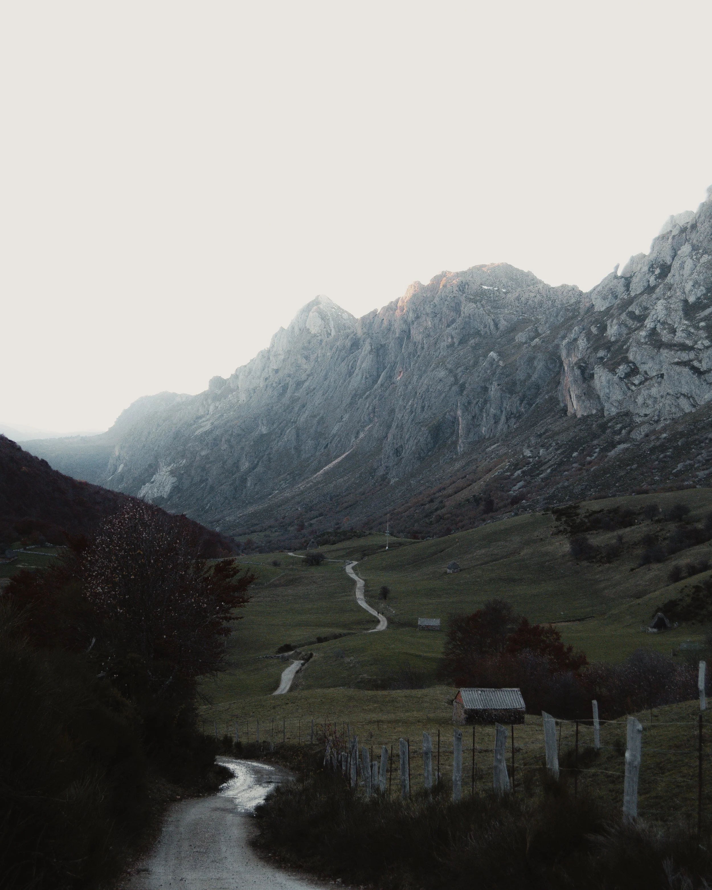 Paisaje de montaña con camino serpenteante, rodeado de praderas y vallado, bajo un cielo despejado.