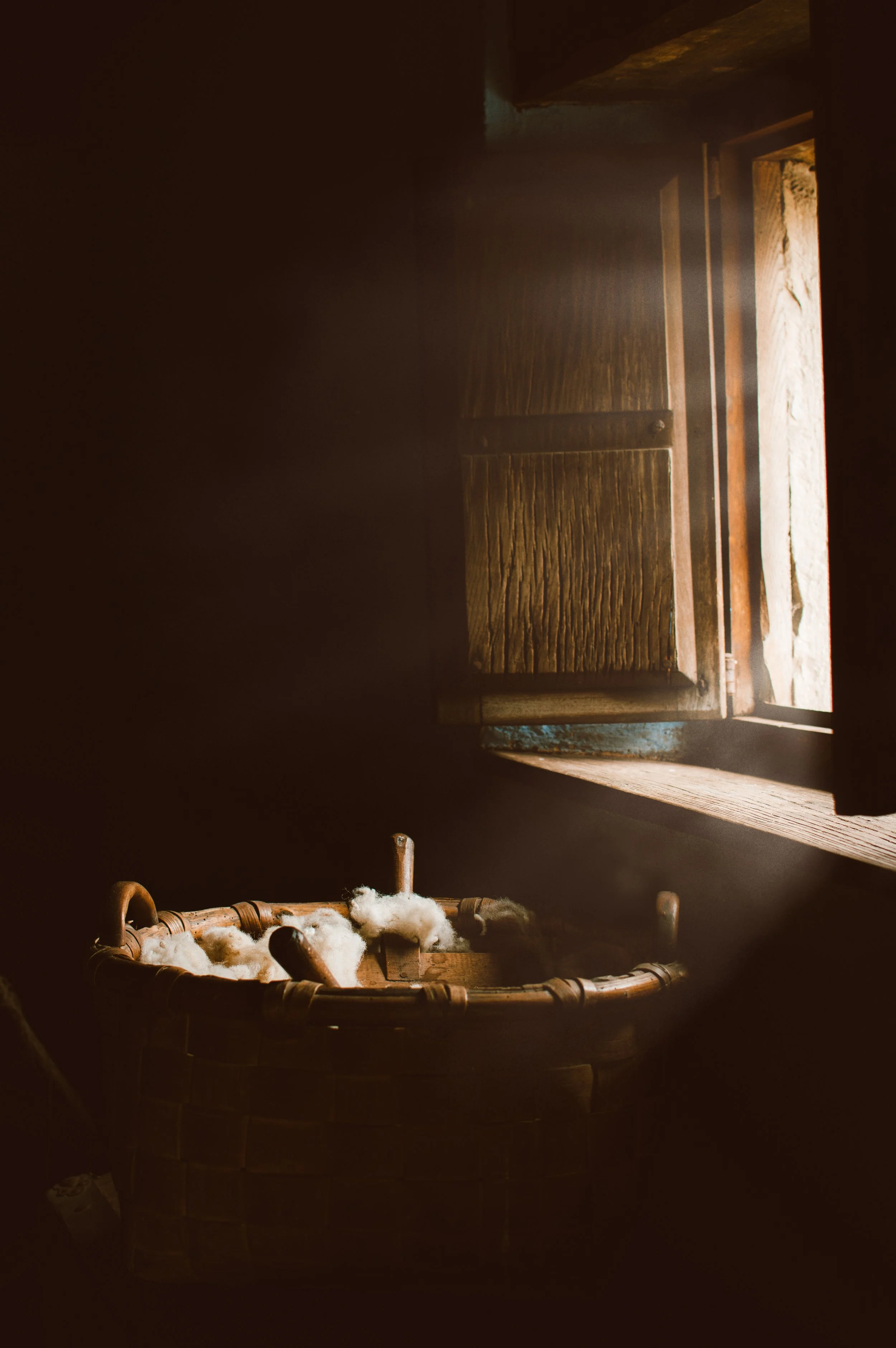 Cesta de mimbre con lana junto a ventana de madera abierta, iluminación cálida.