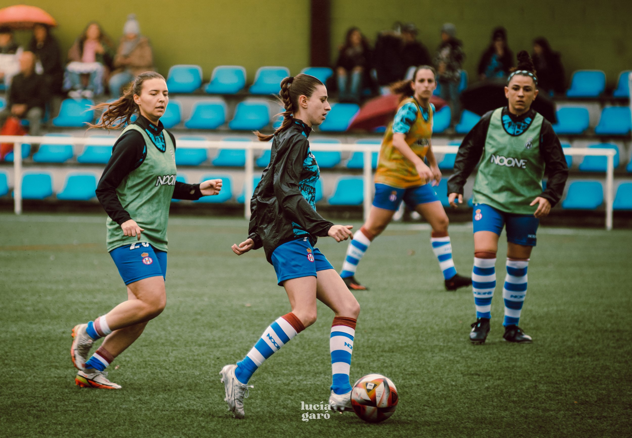 Jugadoras de fútbol entrenando en una cancha; llevan uniforme con medias a rayas y petos de entrenamiento verdes y amarillos. El fondo muestra gradas con asientos azules y espectadores. Una jugadora controla el balón mientras las demás observan.