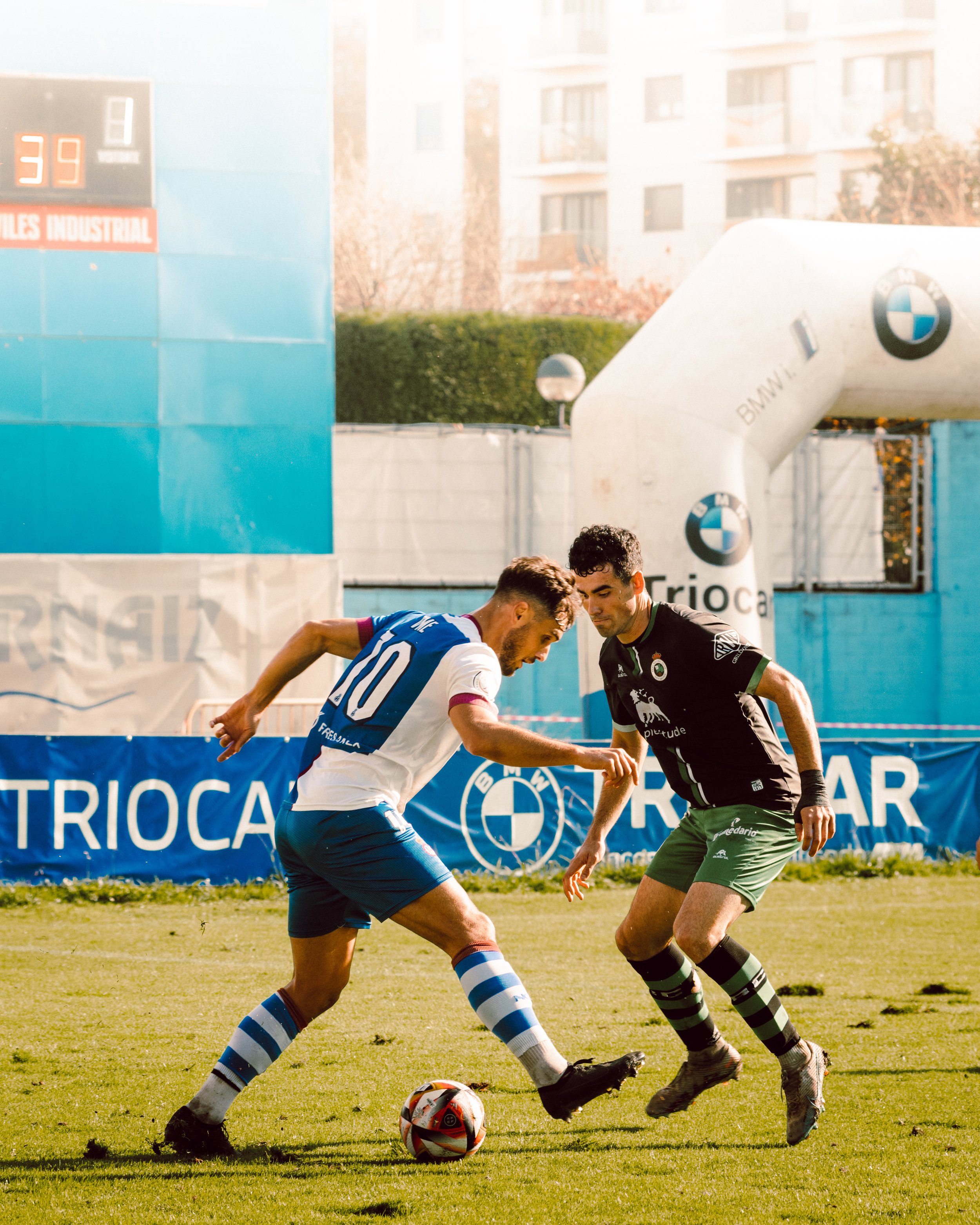 Dos jugadores de fútbol en acción en un partido, uno con uniforme blanco y azul driblando el balón cerca de otro jugador que viste uniforme negro y verde. El marcador y patrocinadores visibles de fondo.
