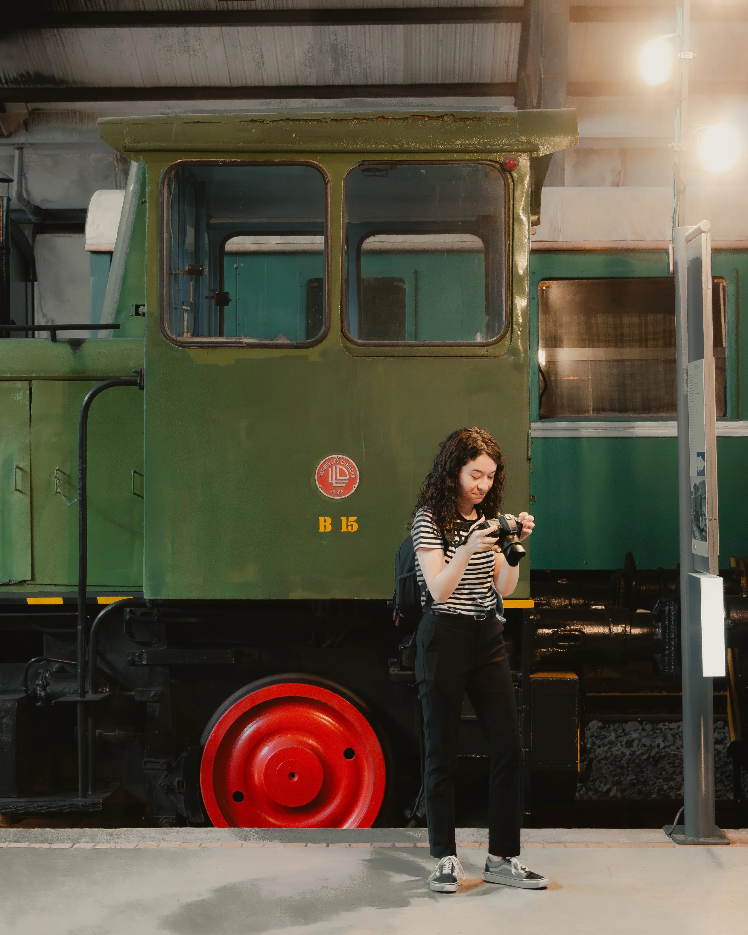 Mujer joven con cámara frente a un vagón de tren verde con una rueda roja, en un entorno interior iluminado.