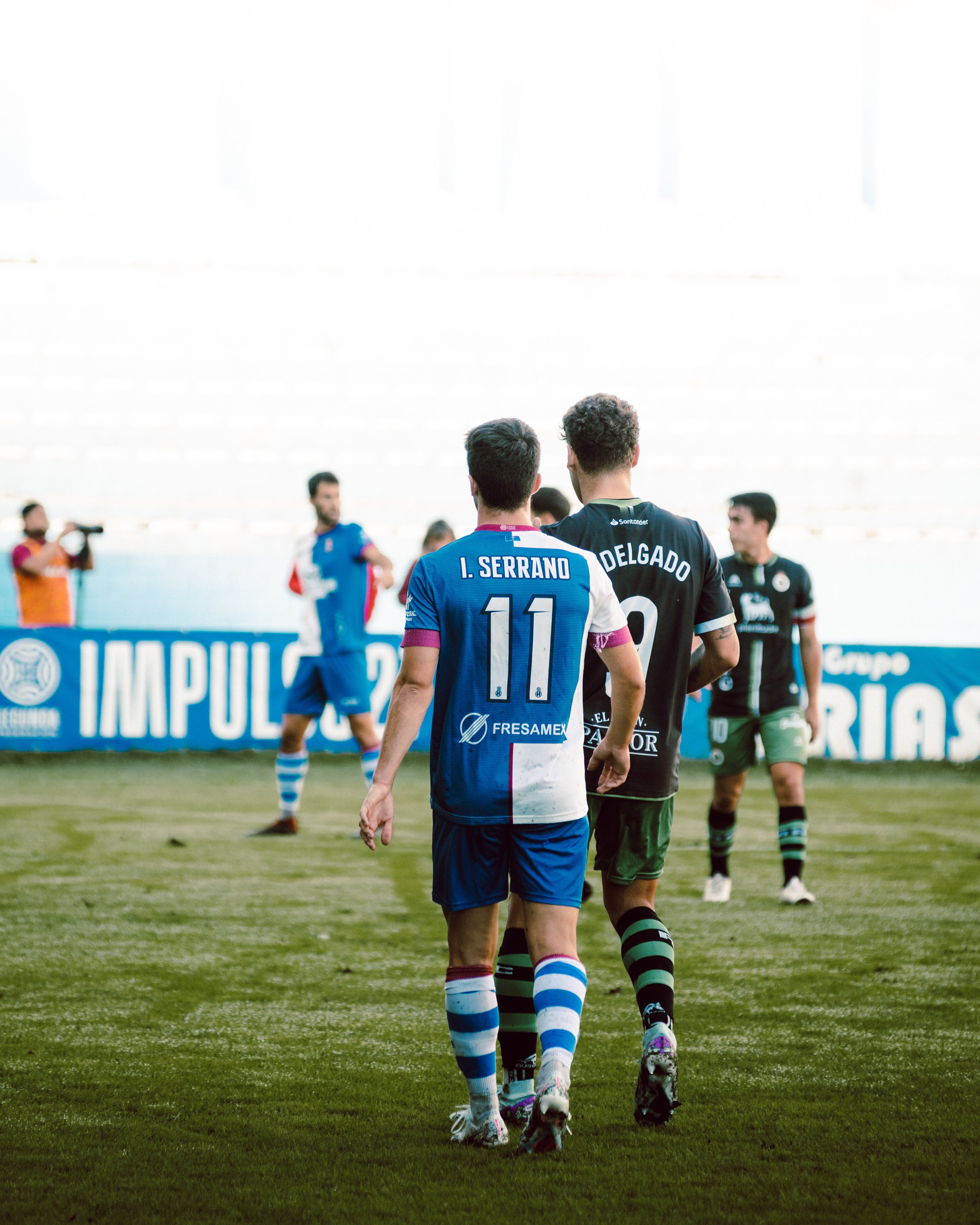Jugadores de fútbol en el campo, uno con camiseta azul y blanca, otro con camiseta negra y verde, vistos de espaldas. Publicidad visible en el fondo.