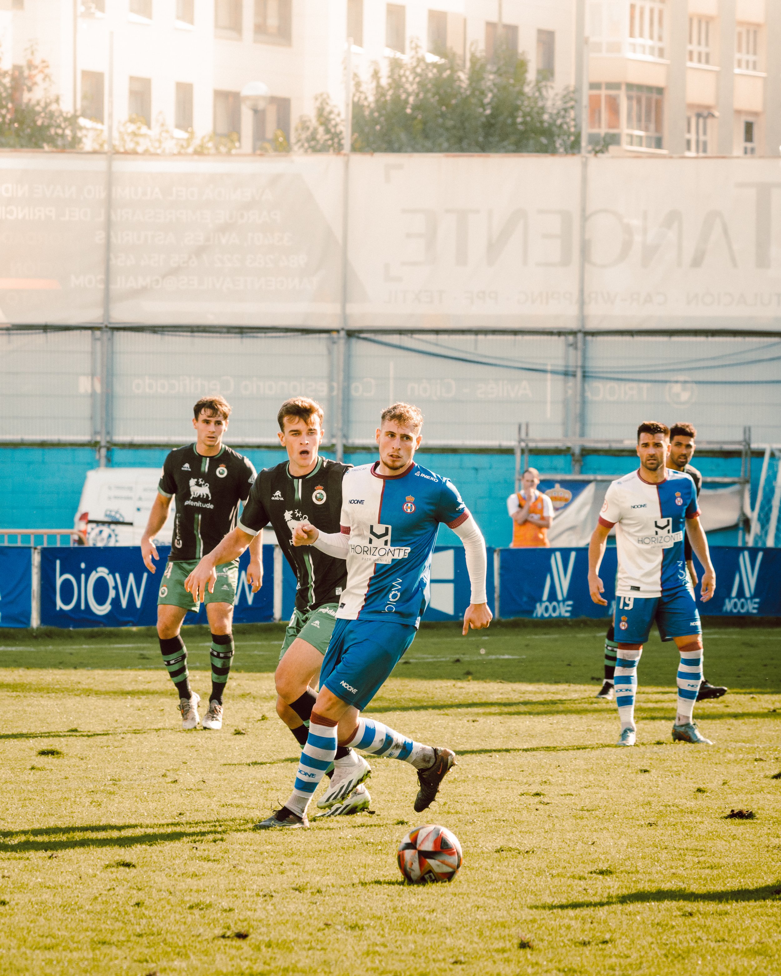 Partido de fútbol con jugadores en acción, uno de ellos en uniforme azul y blanco controlando el balón.
