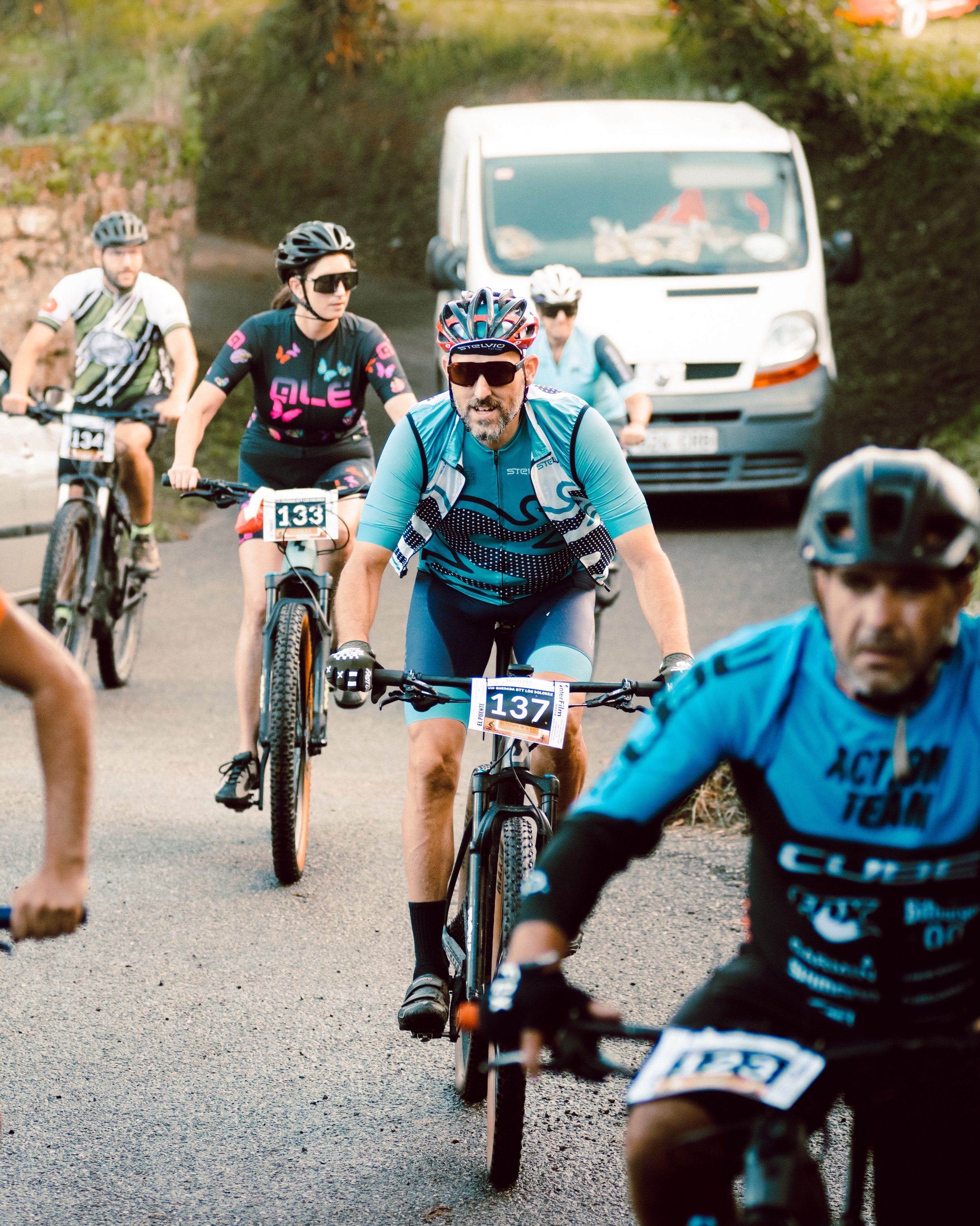 Grupo de ciclistas participando en una carrera, usando ropa deportiva y cascos. Al fondo, un vehículo blanco estacionado a un lado del camino.
