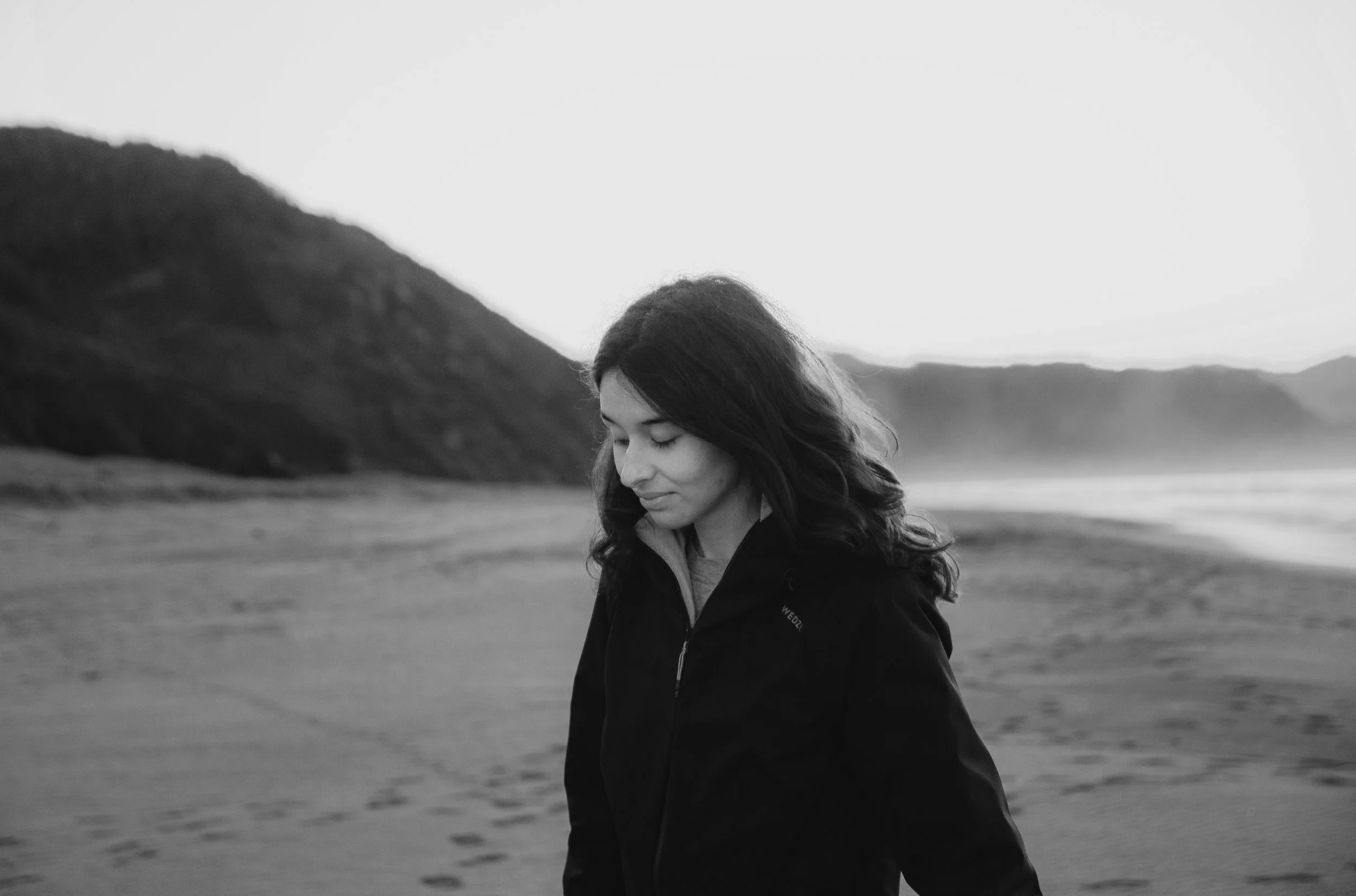Mujer caminando en la playa, paisaje montañoso de fondo. Imagen en blanco y negro.