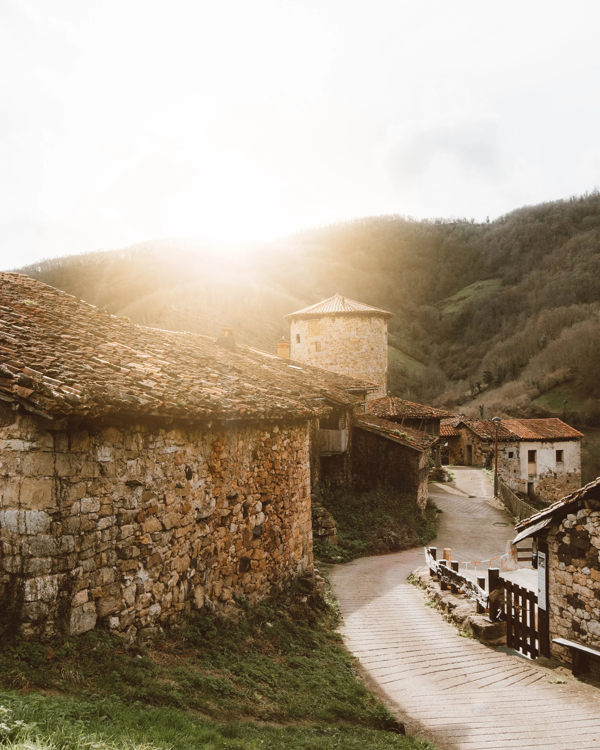 Pueblo rural con casas de piedra y techos de tejas, camino curvo, paisaje montañoso al fondo, luz del sol intensa. en Bandujo, Asturias.