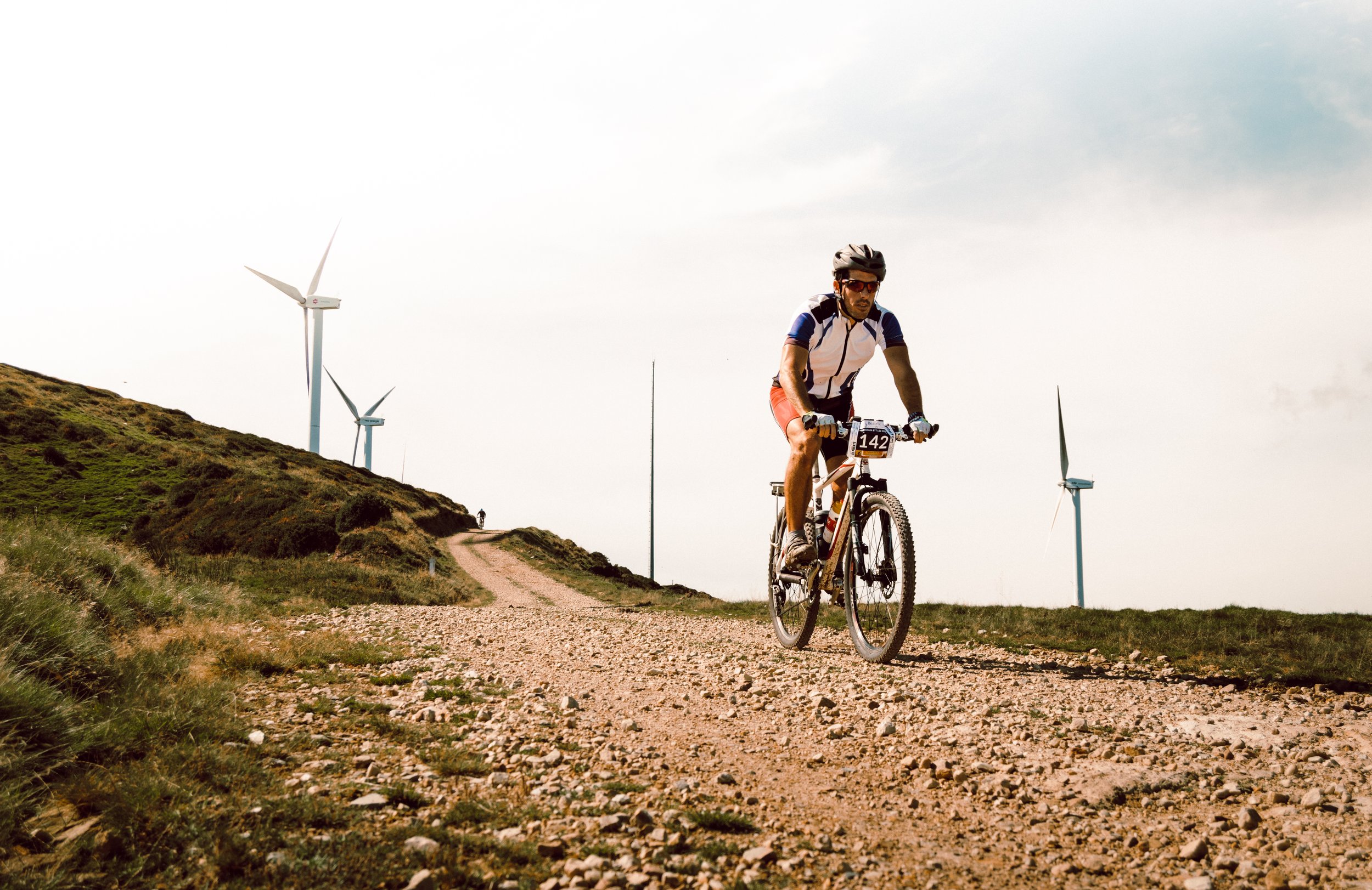 Ciclista compitiendo en una carrera de ciclismo de montaña frente a aerogeneradores en un paisaje rural.