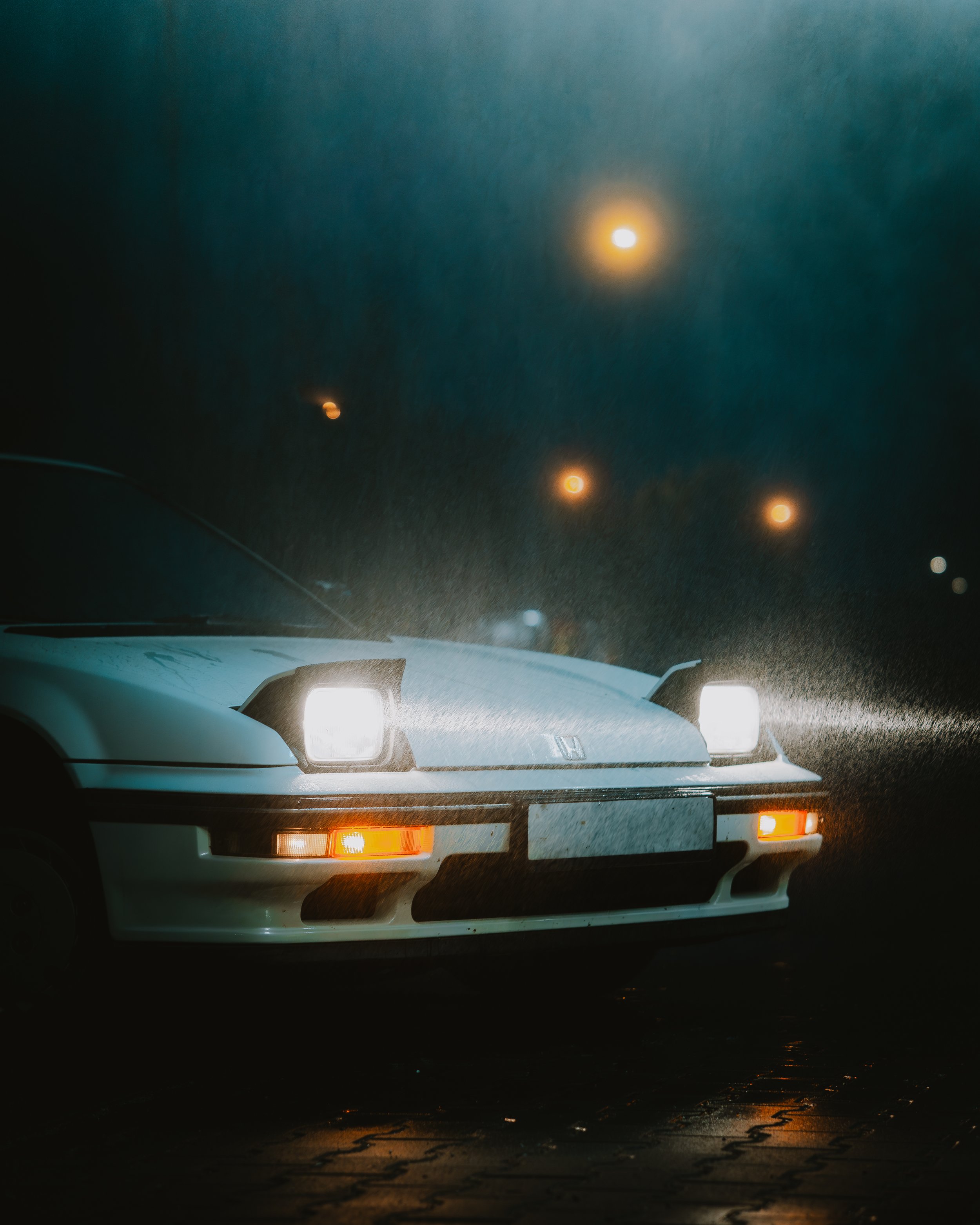 Coche blanco en una calle bajo la lluvia, con luces delanteras encendidas en la noche.