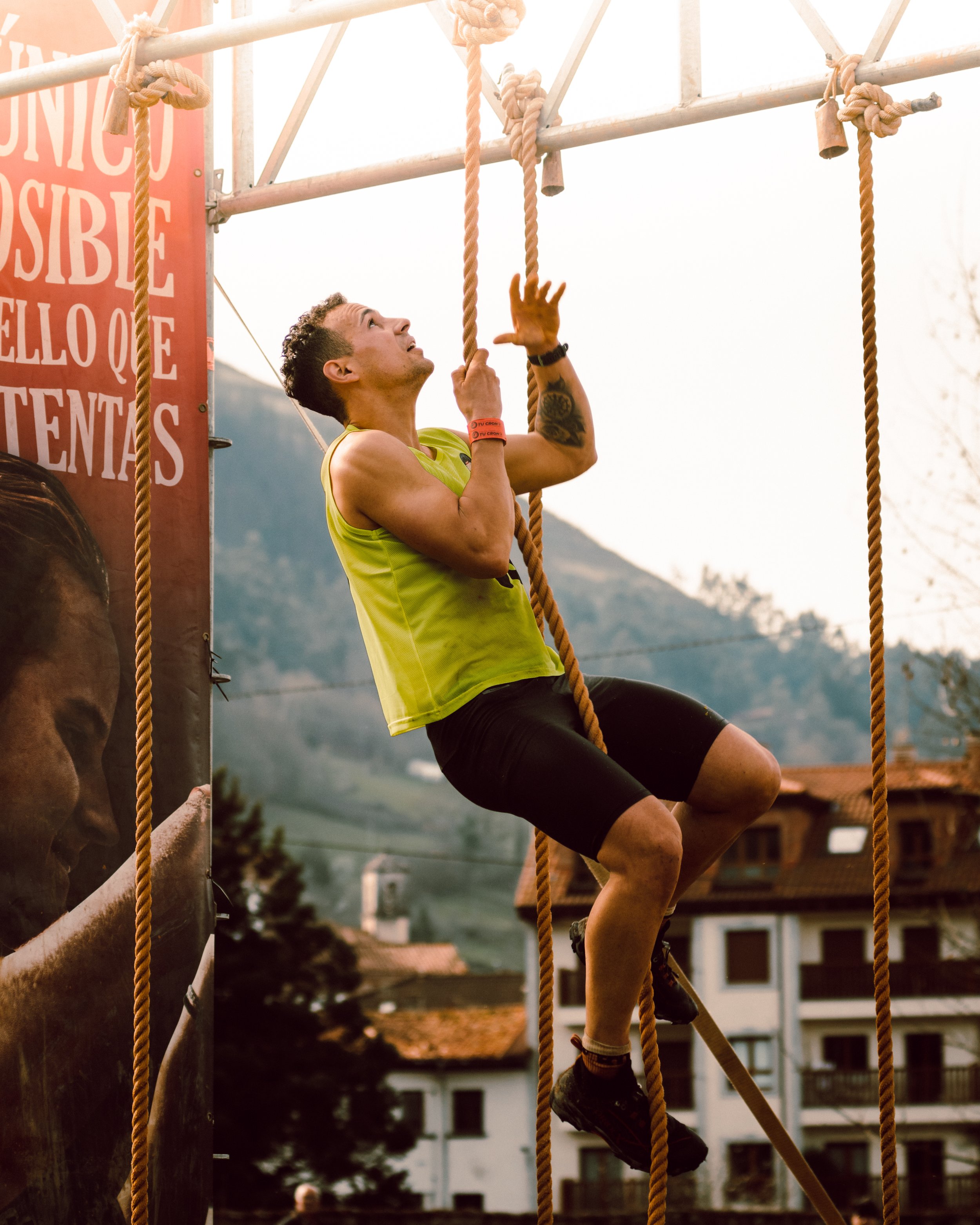 Hombre subiendo una cuerda en una competición al aire libre, con edificaciones y montañas de fondo.