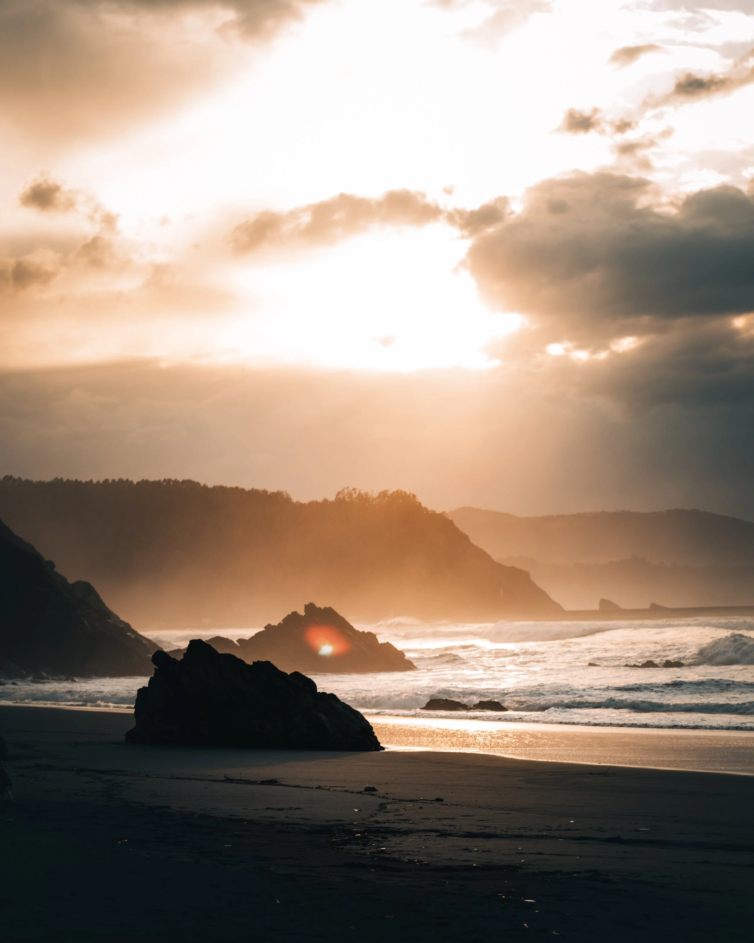 Playa al atardecer con nubes, rocas en la arena y olas del mar.