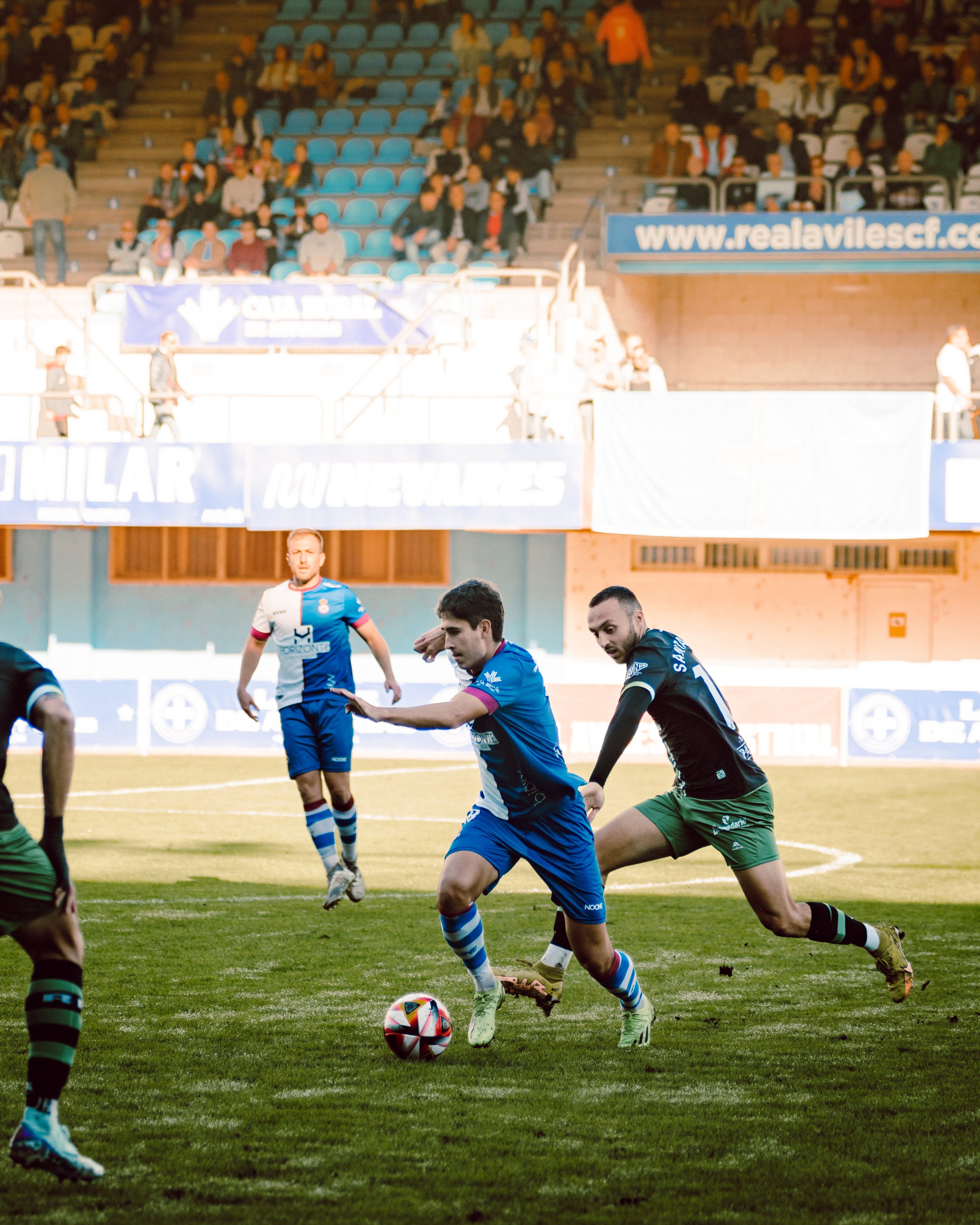 Jugadores de fútbol compitiendo en un partido en un estadio, con espectadores en las gradas.