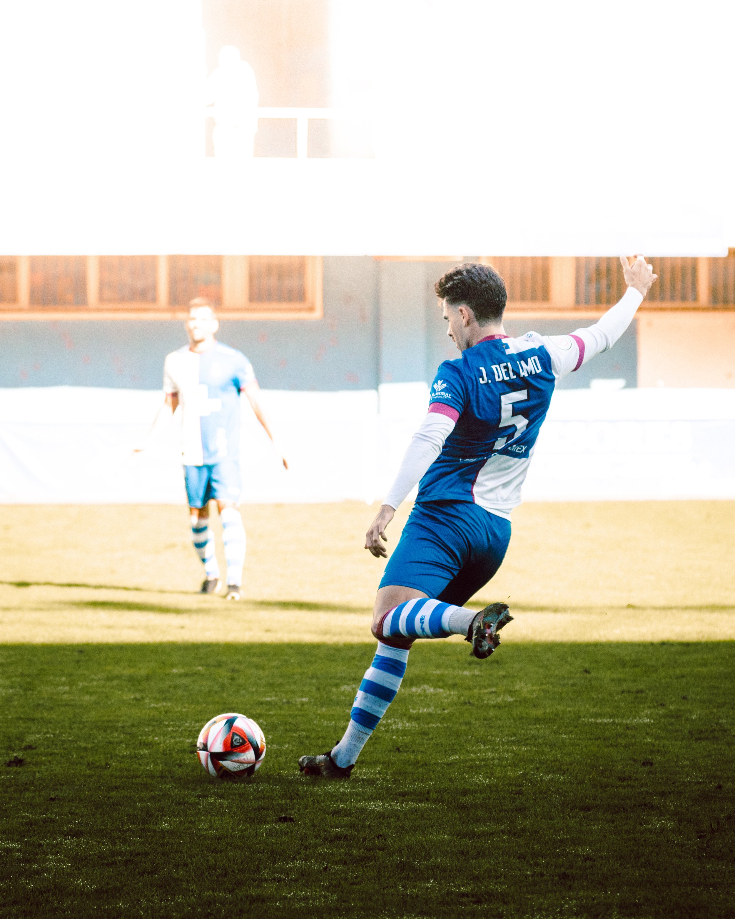 Futbolista ejecutando un tiro en un campo de fútbol, con uniforme azul y blanco. Otro jugador está de fondo.