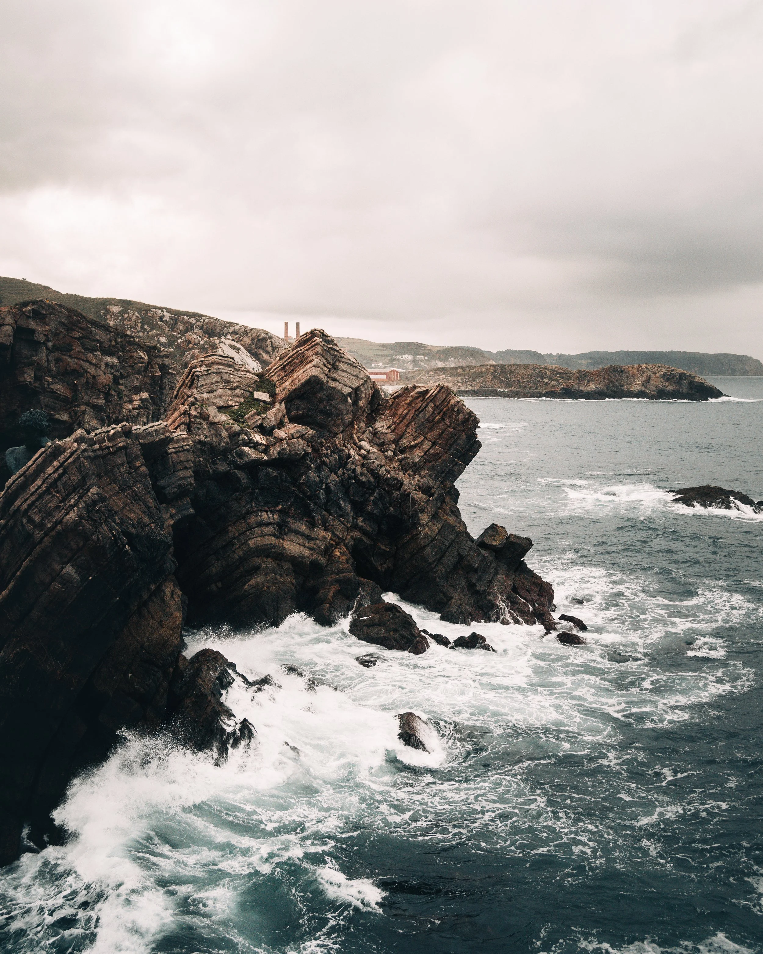 Costa rocosa con acantilados inclinados, olas rompiendo en las rocas y un cielo nublado.