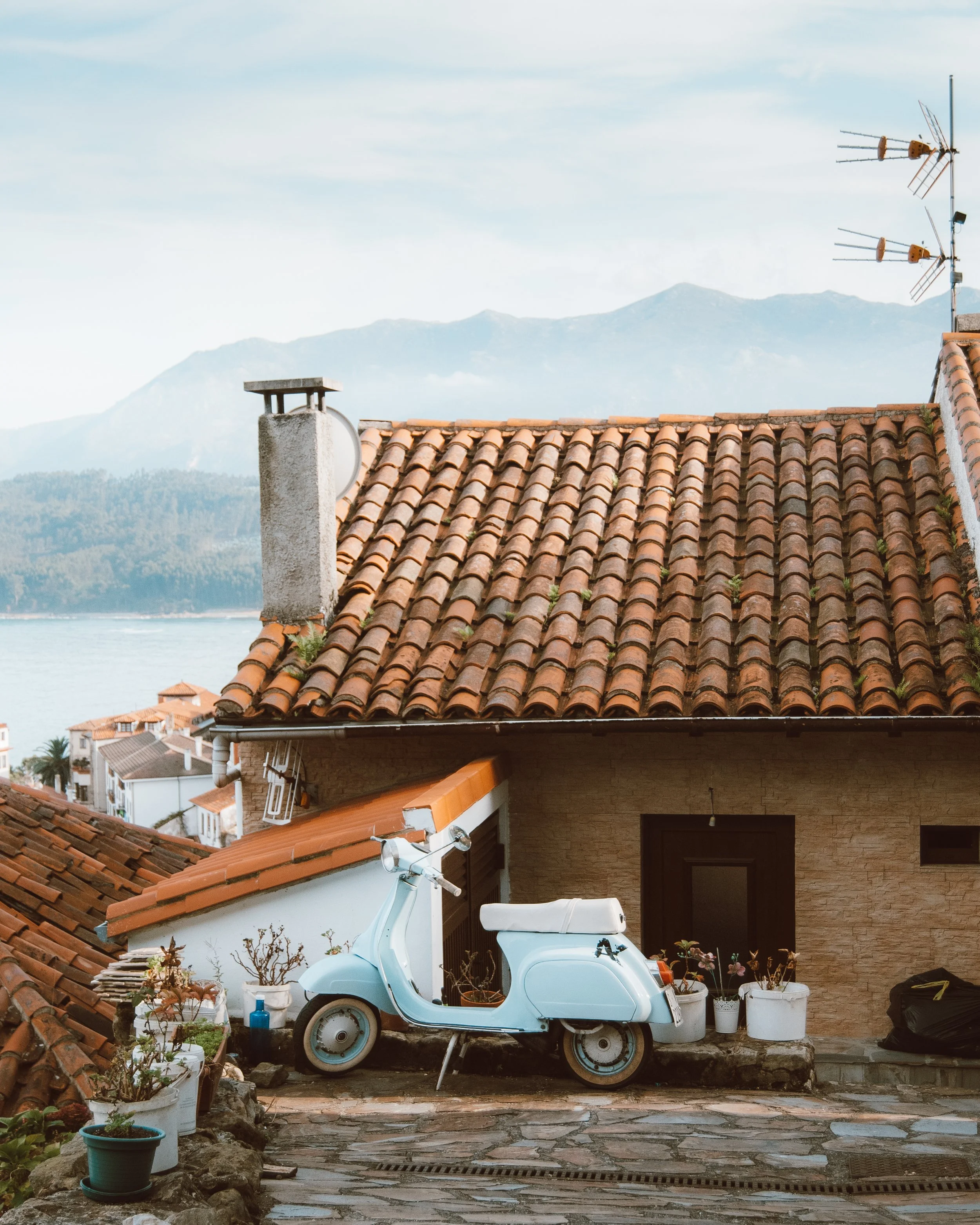 Motocicleta tipo Vespa blanca estacionada frente a una casa con techo de tejas rojas y macetas, con vista a las montañas y un lago al fondo.