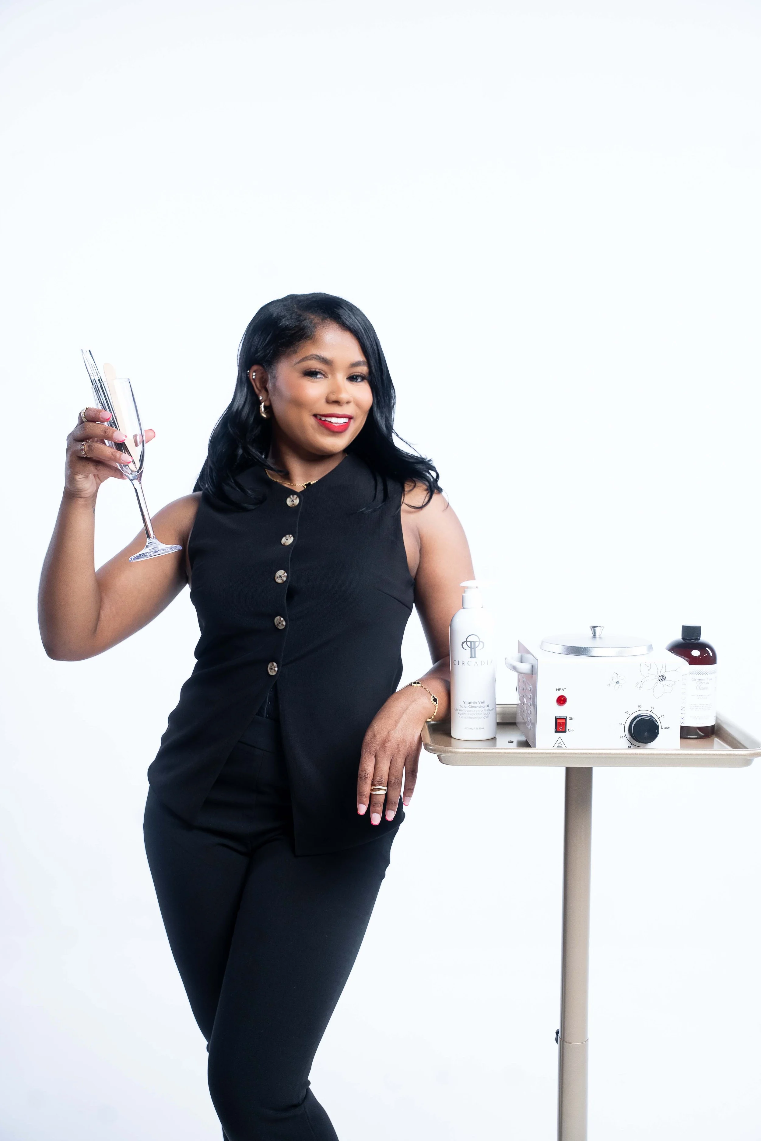 Woman with black hair and red lipstick holding a champagne flute, standing next to a medical skincare device on a side table with bottles and supplies.