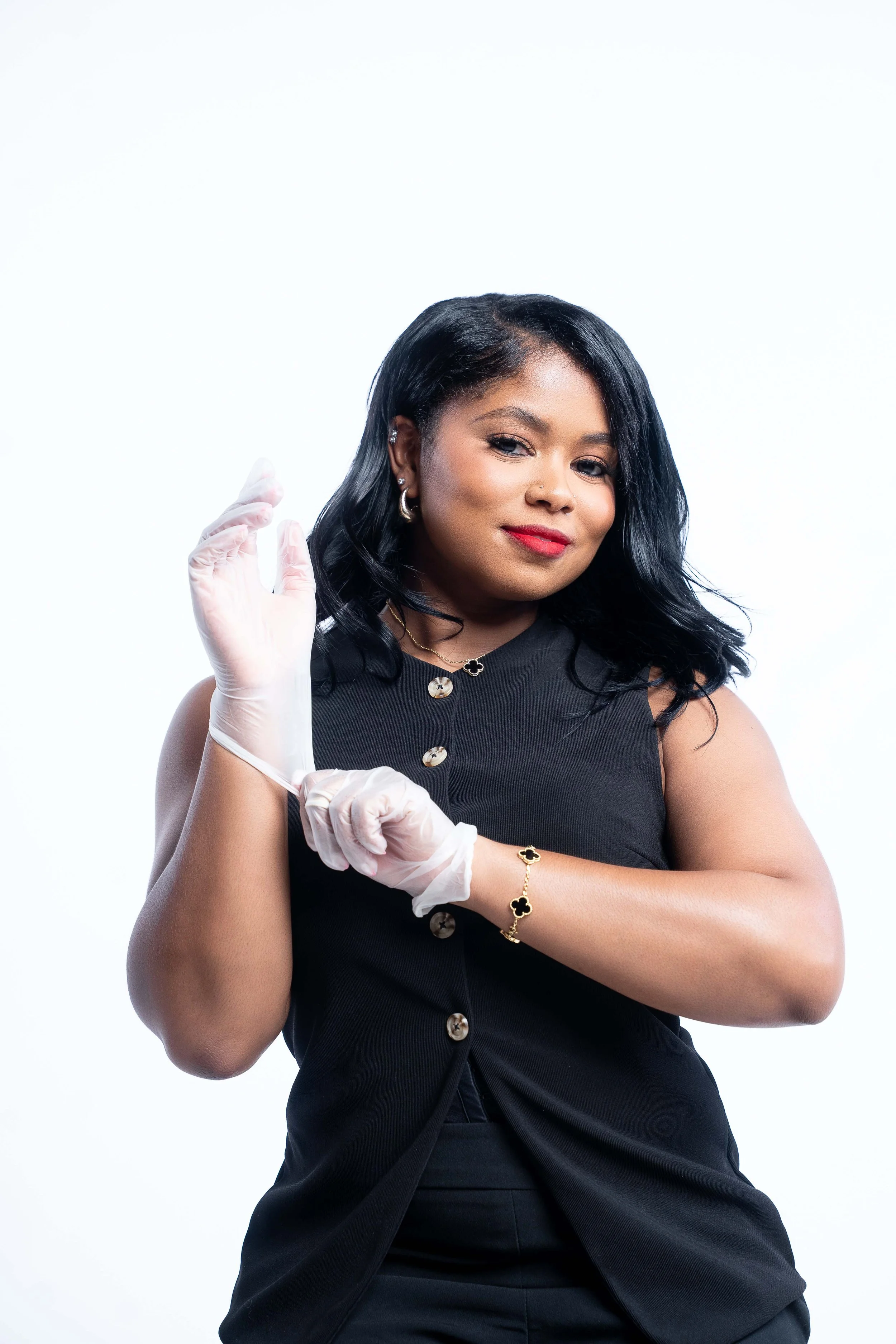 A woman with black hair in a black sleeveless top, wearing gold jewelry, and putting on white gloves, standing against a plain white background.