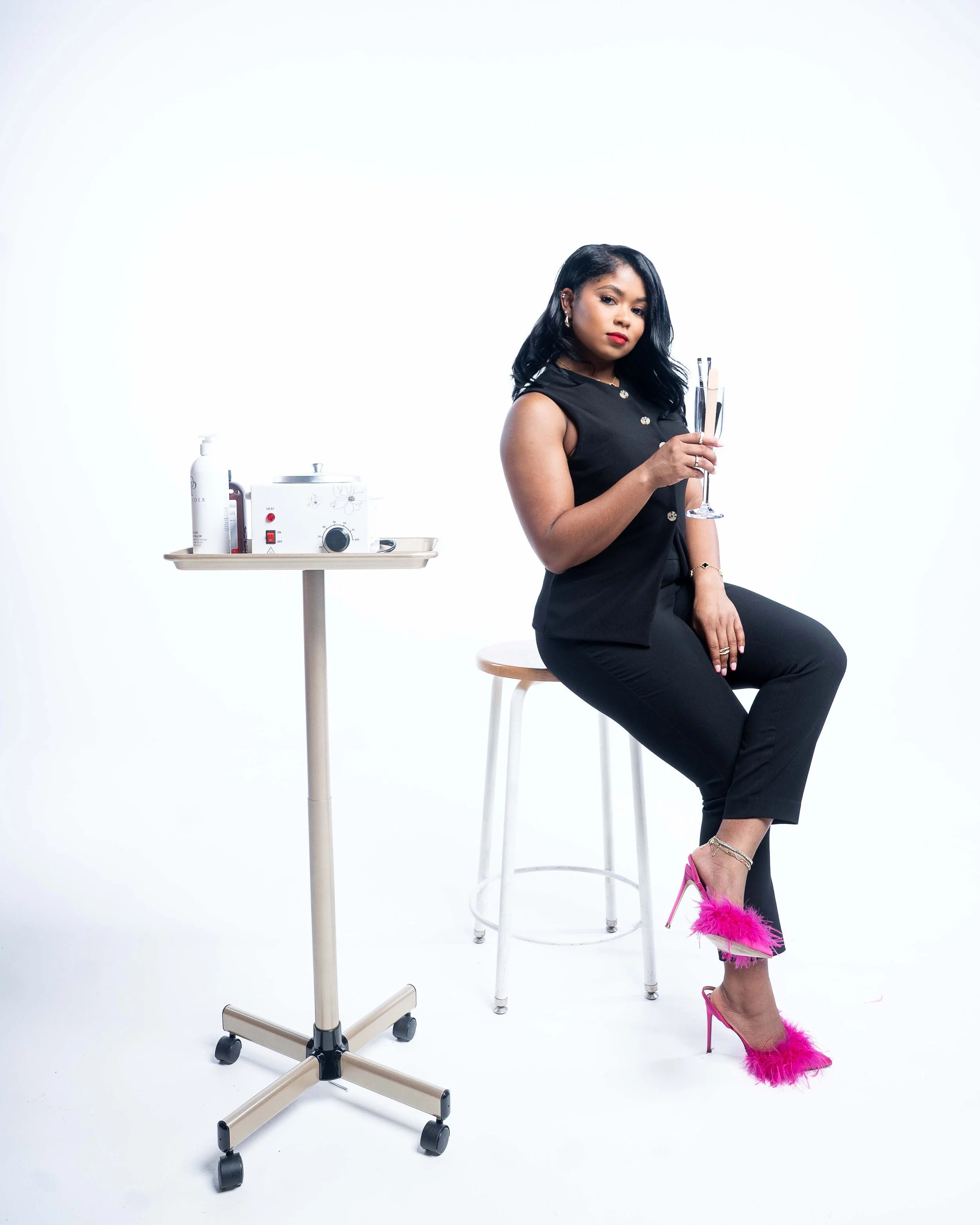 A woman dressed in black high heels with pink feathers, black pants, and a black sleeveless top, sitting on a stool while holding a glass of champagne in a studio with white background. There is a small medical or skincare device on a stand to her left with bottles.