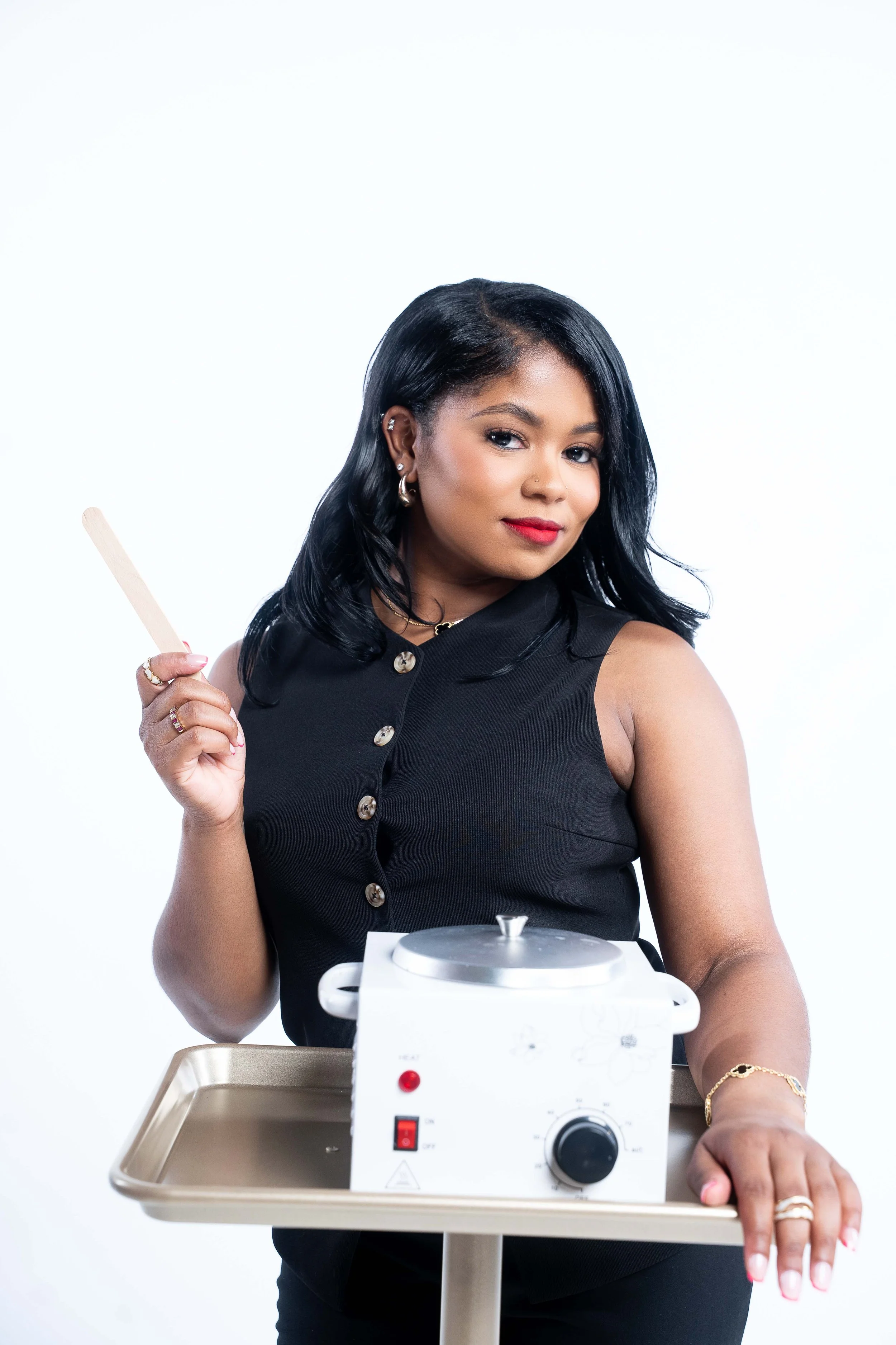 A woman with black hair, wearing a black sleeveless top, holding a heat paddle, standing next to a small white device on a beige tray, set against a plain white background.