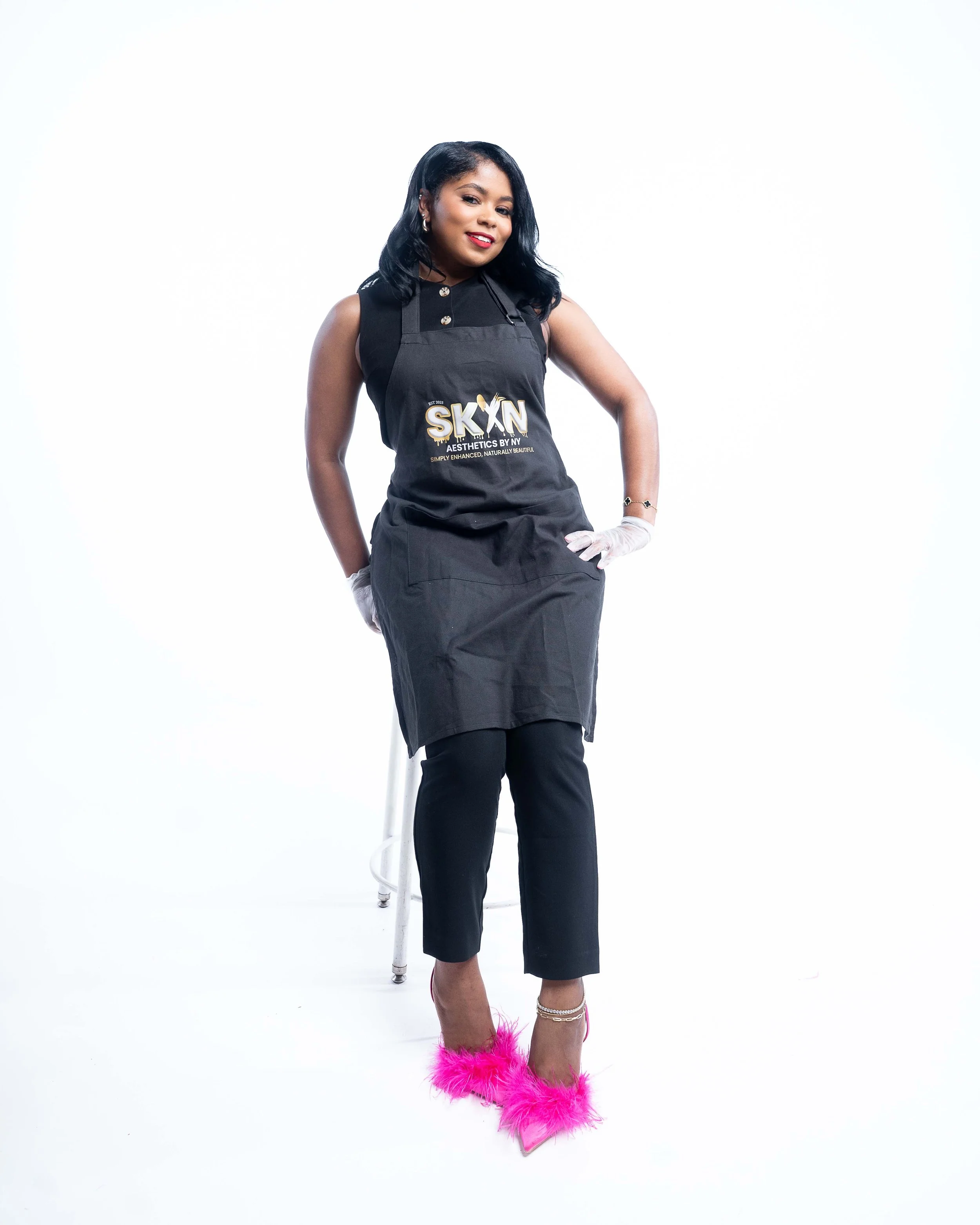 A woman in a black apron and black pants standing against a white background, wearing pink feathered heels and smiling at the camera.