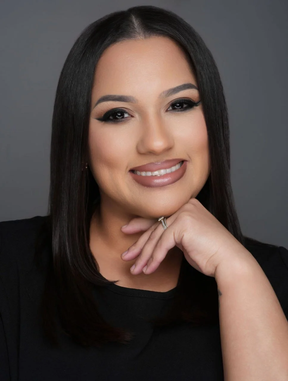 A woman with long dark hair, makeup, and a ring, smiling and resting her chin on her hand against a gray background.