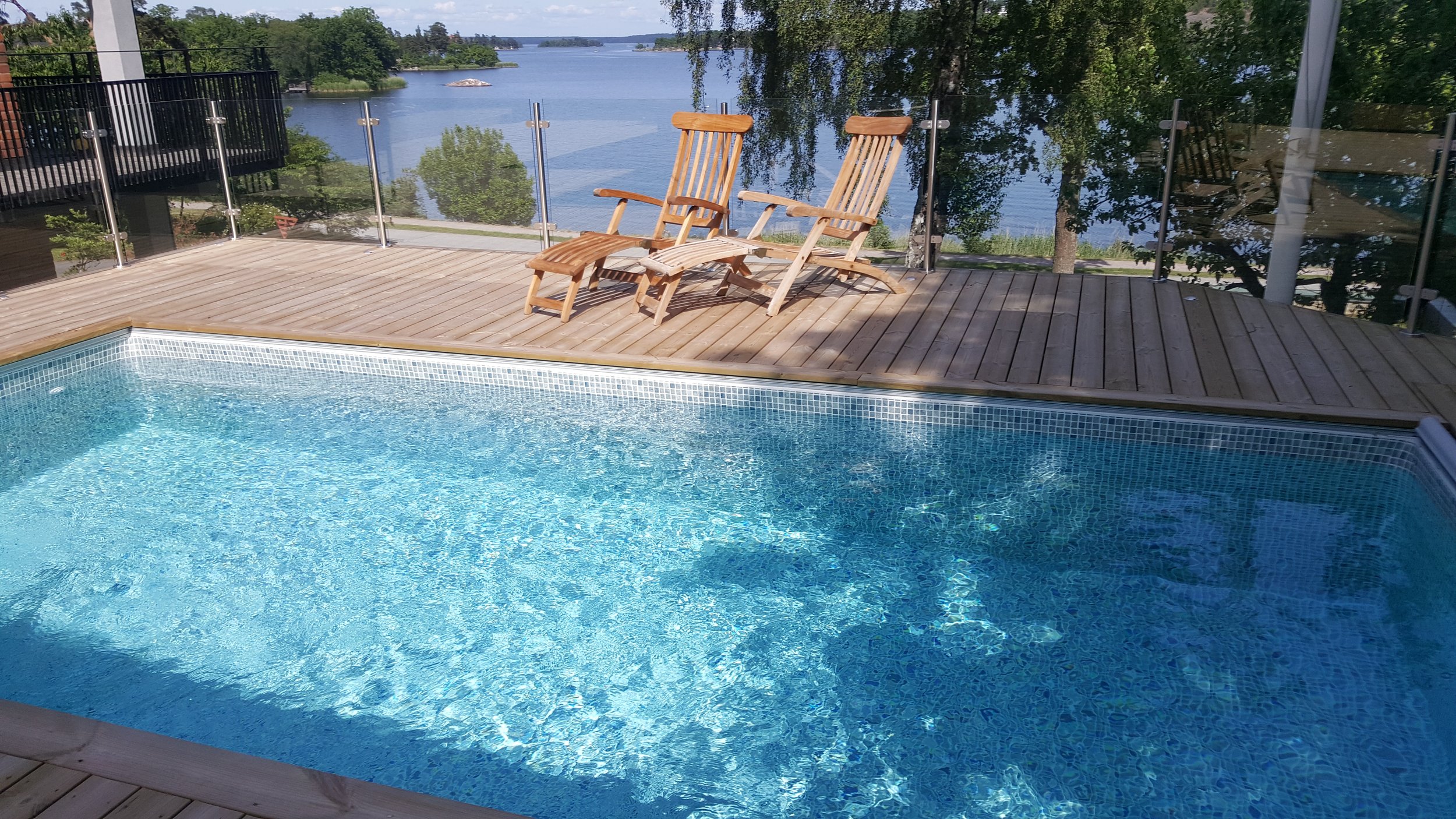 Poolområde med trädäck och två solstolar framför en sjö.