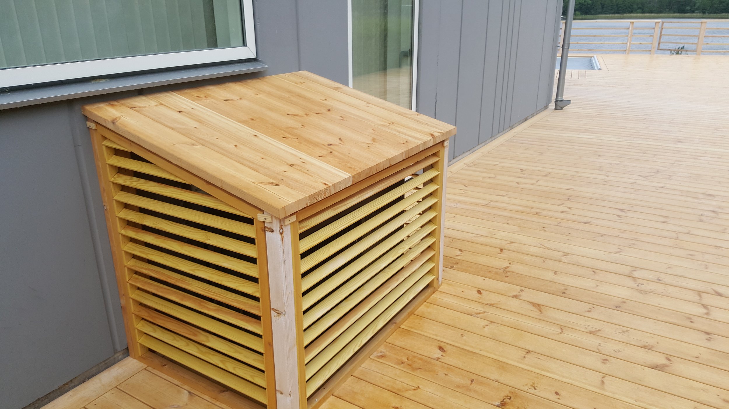 Utomhus AC-kompressor täckt med träribbor, placerad på en träaltan intill en byggnad med fönster.