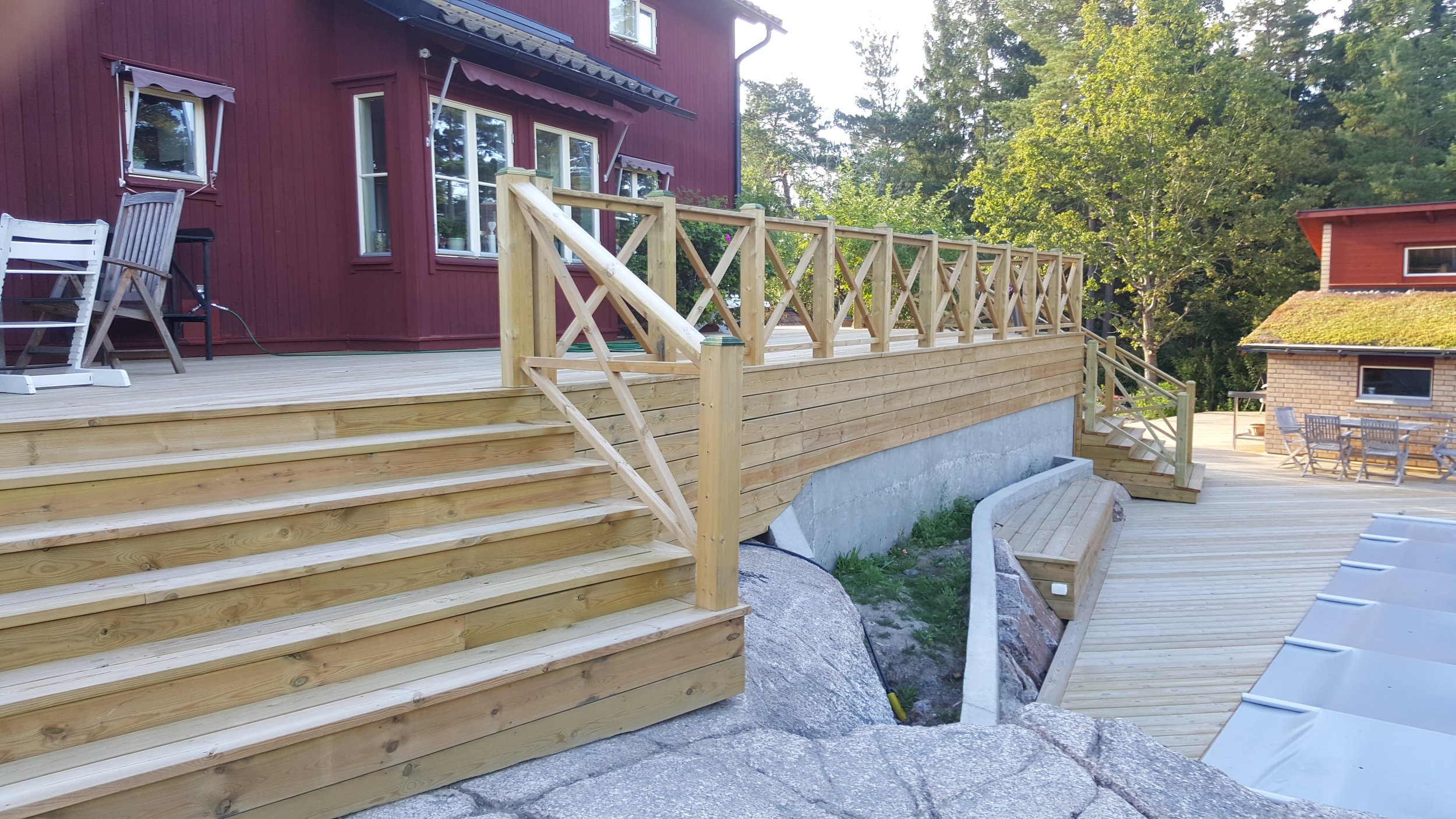 Två röda hus med träveranda och trappa i trä, omgivna av träd.