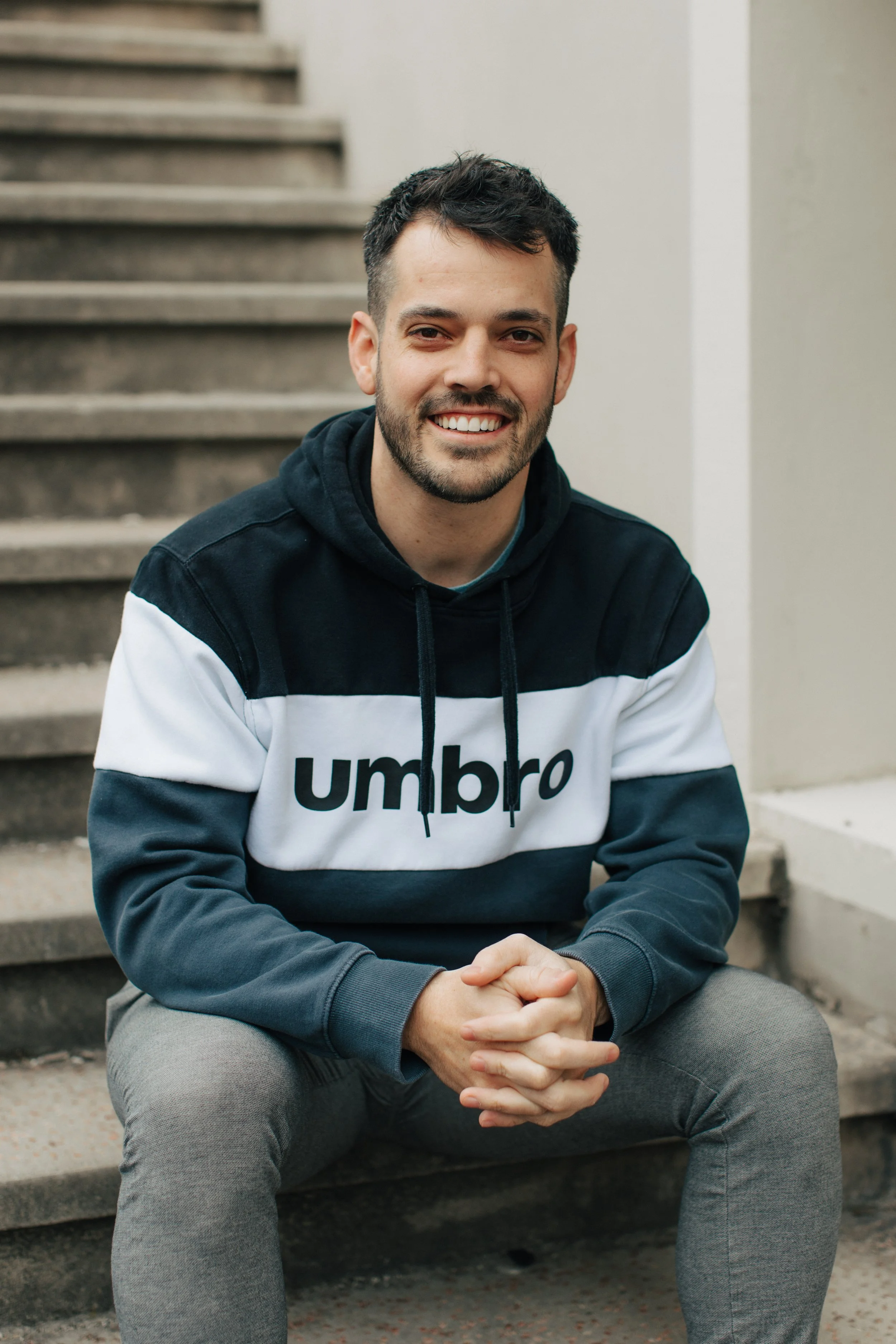 Junger Mann sitzt lächelnd auf Treppe, trägt schwarzen und weißen Hoodie mit 'Umbro' Logo