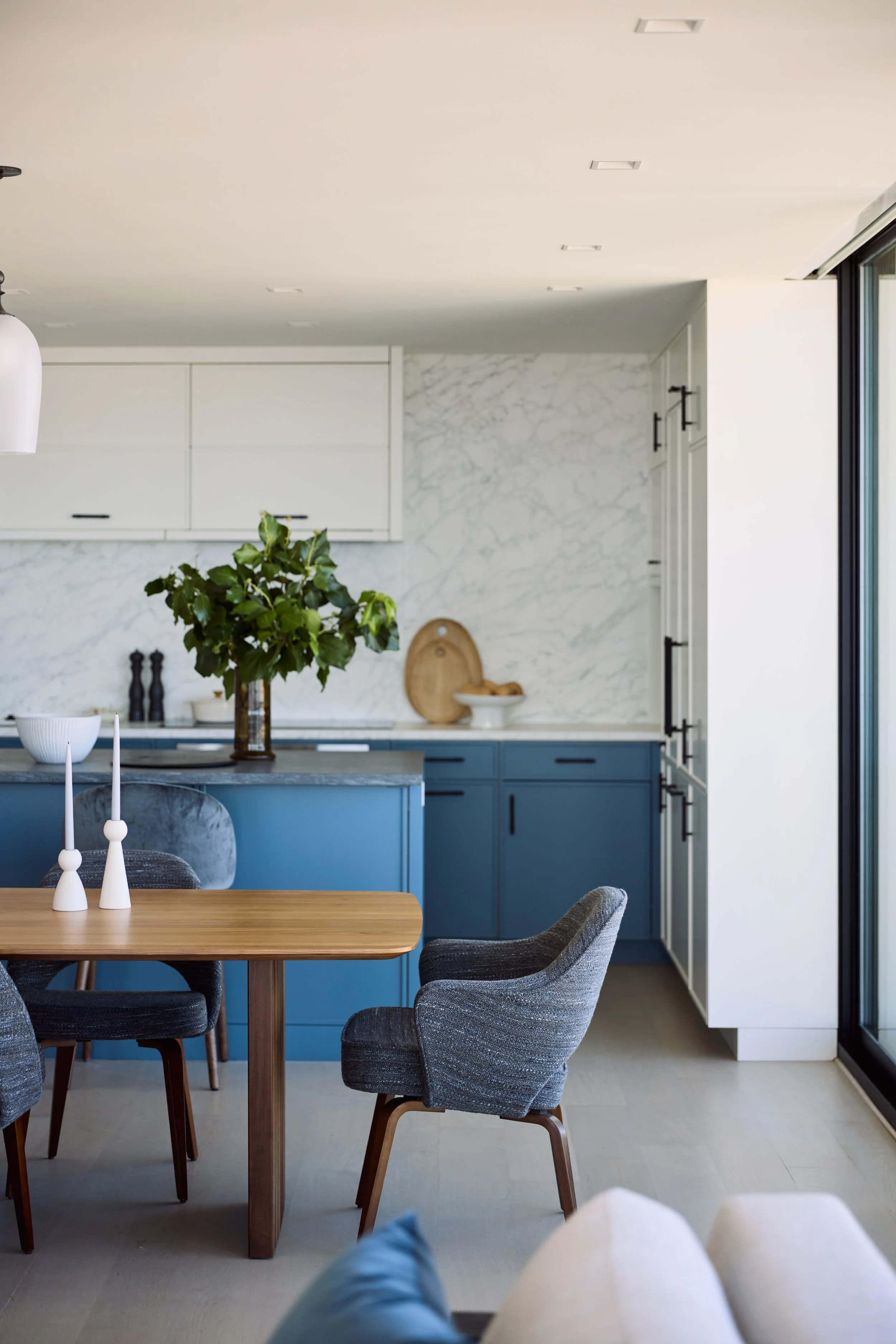 Modern kitchen with blue cabinets, marble backsplash, and a wooden dining table with gray upholstered chairs. Decor includes white candlesticks and greenery, with sliding glass doors on the right.