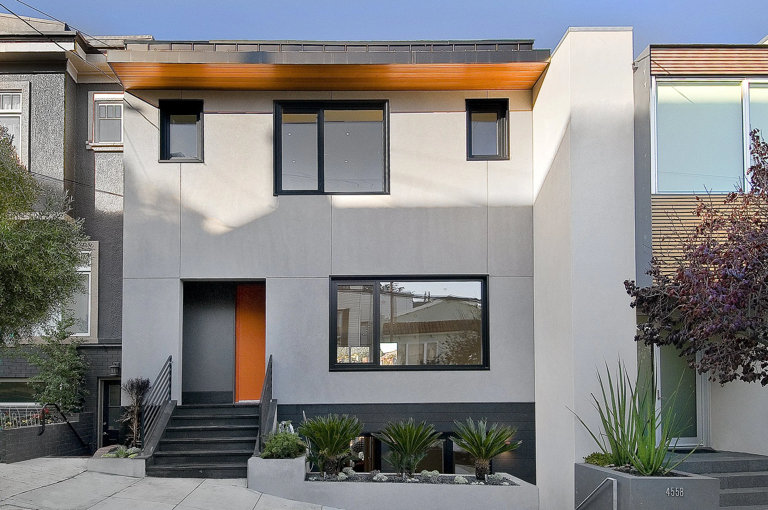 Modern multi-story house with white exterior, black-framed windows, an orange front door, front garden with plants, stairs leading to the entrance, and neighboring buildings. Passive home with sustainable features project in San Francisco, Ca.