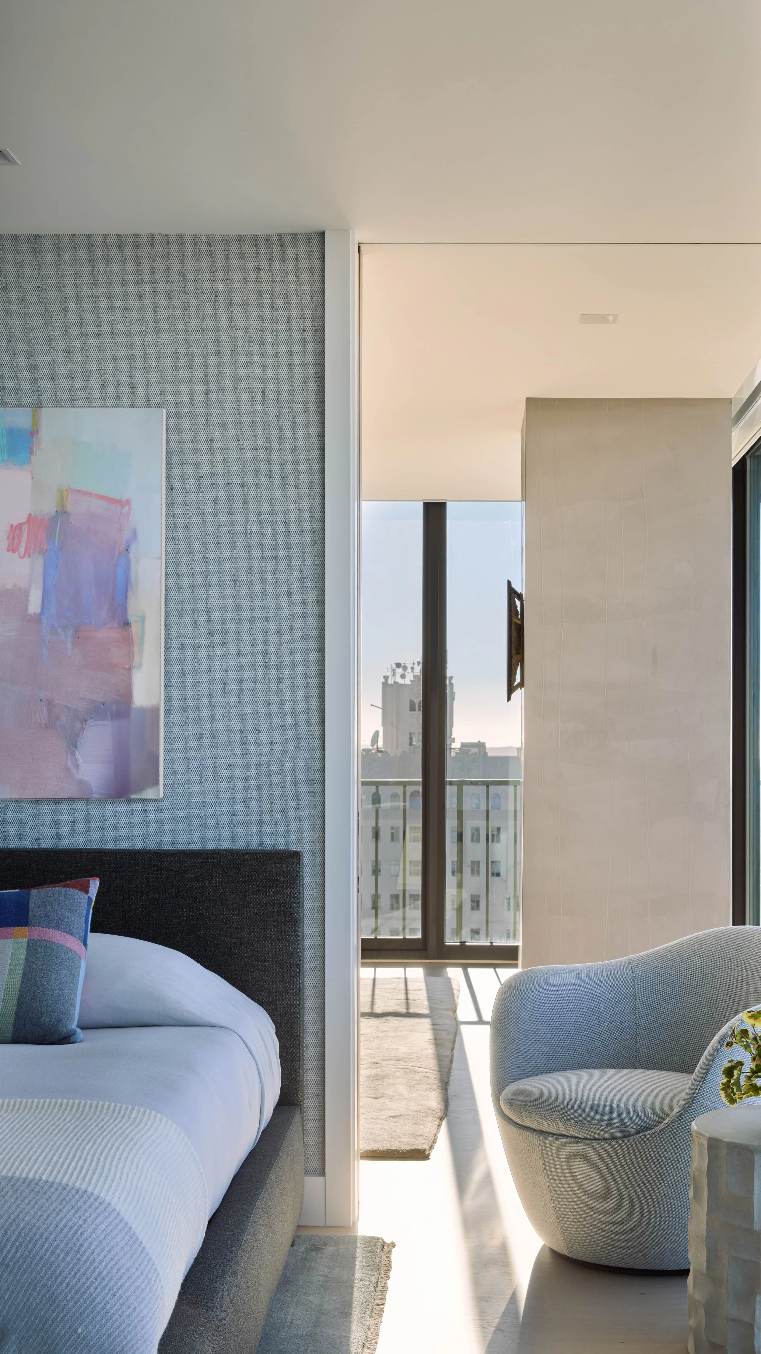 Modern bedroom with a dark upholstered bed, white bedding, colorful pillow, abstract art on the wall, and a round light gray armchair near a large glass door leading to a balcony.