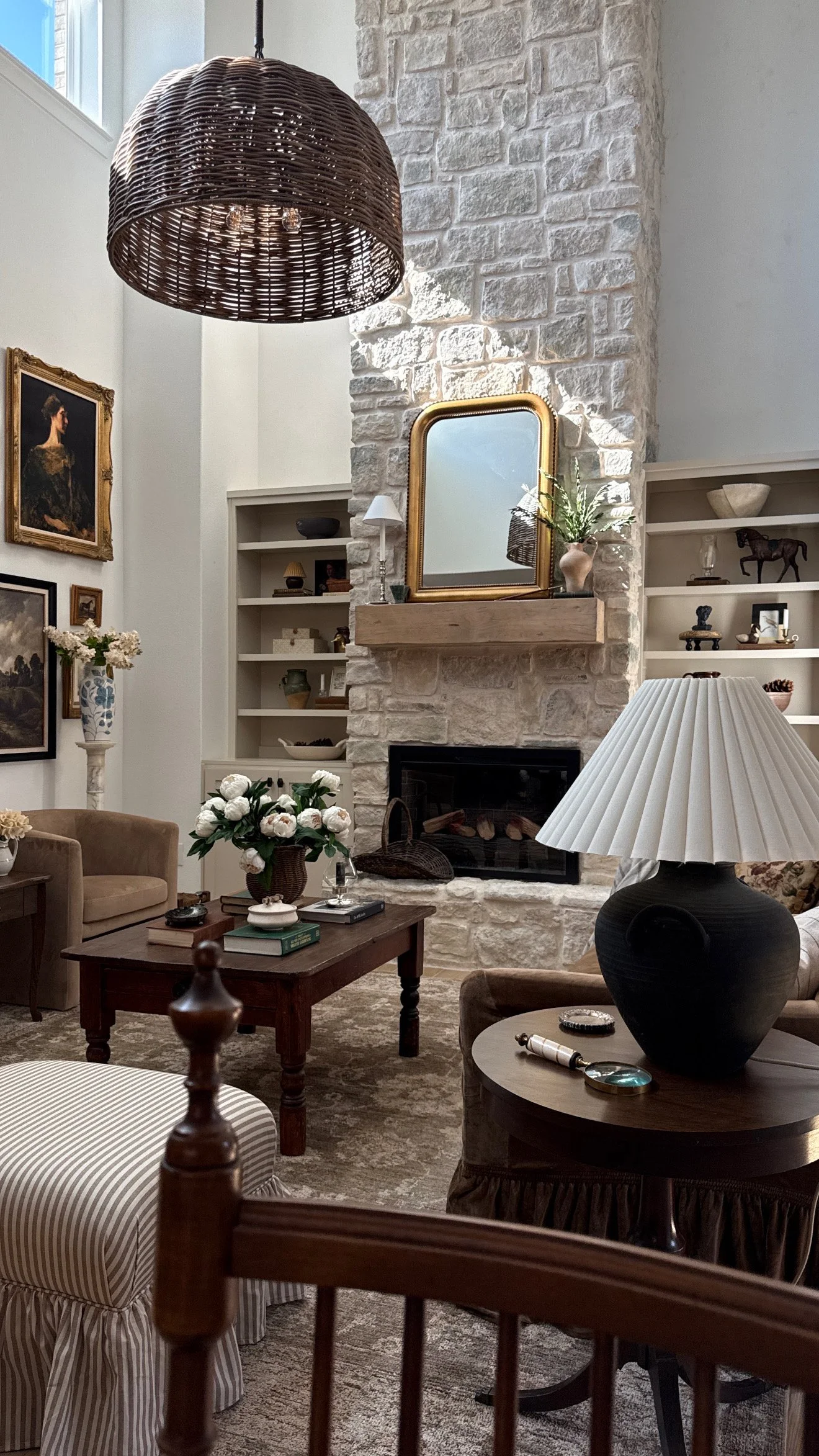 A cozy living room with a stone fireplace, white built-in shelves, framed artwork, a mirror, a bouquet of white flowers, and various decorative items, with a table lamp and furniture.